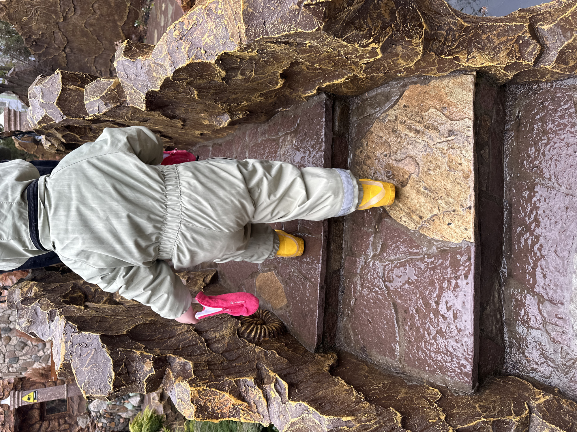 A child wearing a tan jumpsuit and yellow shoes is walking on a rough, textured, brown and gold surface that appears to be part of a stone structure. The child is also wearing pink mittens. The surface is adjacent to a wet, brown stone basin. The scene appears to be outdoors. The structure looks like a decorative rock feature near a pond. Based on the setting, it could be within Gorky Park in Izhevsk, Russia.