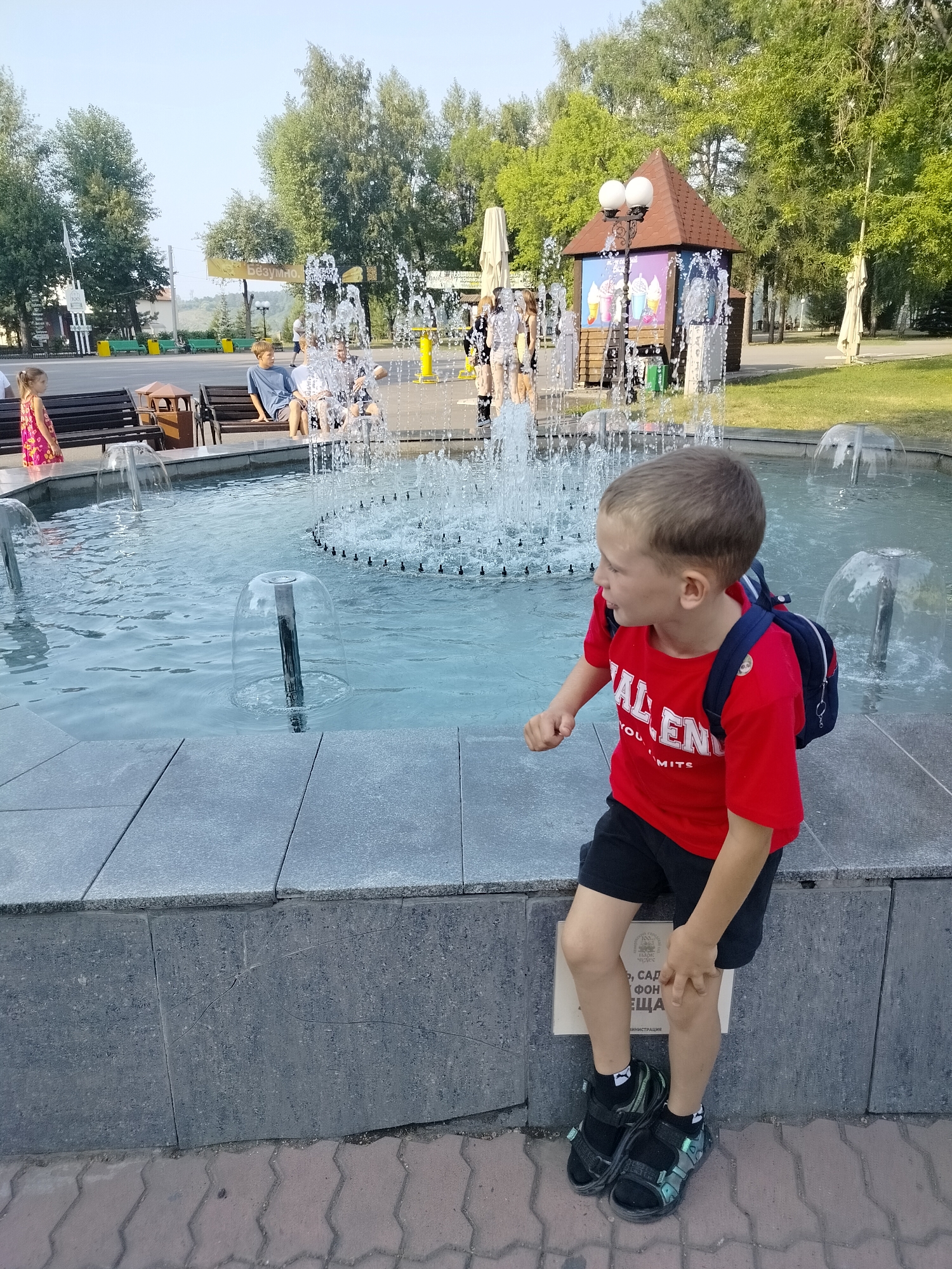 The image shows a boy wearing a red t-shirt, black shorts, and sandals, with a backpack, sitting on the edge of a fountain in Kemerovo, Russia. He is looking to his right. In the background, there are other people, trees, a small building with an advertisement for ice cream, and more fountains. The fountain's water is clear and sparkling. The ground around the fountain is paved with gray stone and brick. The sky is clear and blue.
