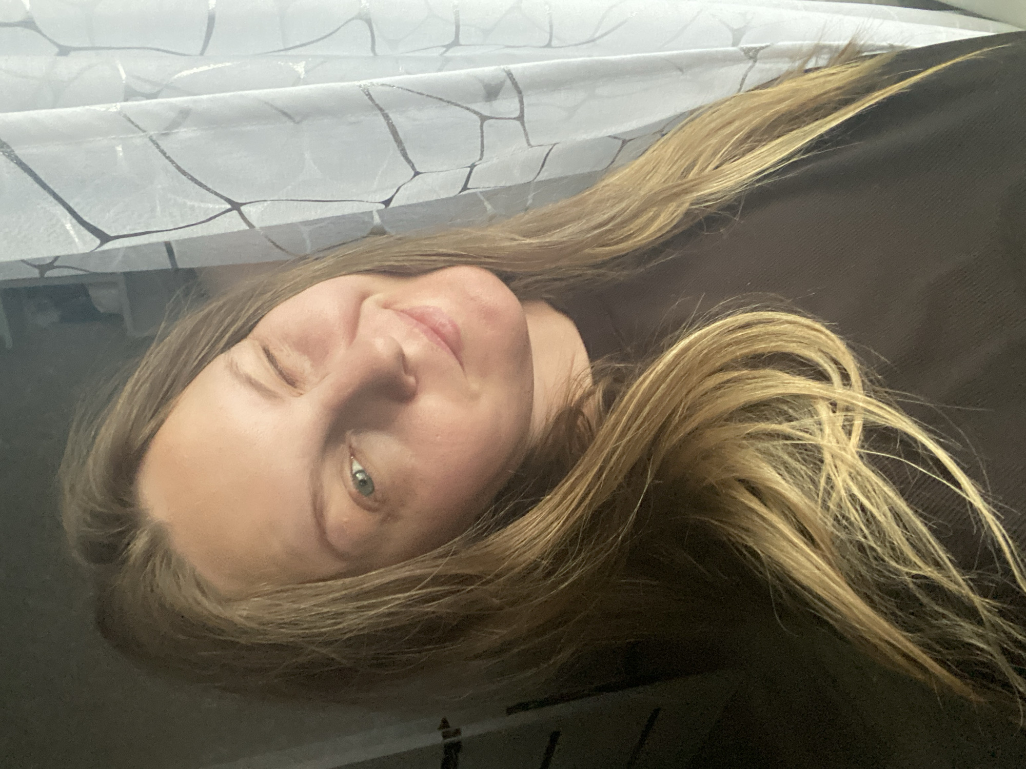 A young woman with blonde hair is lying down, her head tilted to the side, winking. She is wearing a dark brown top. A sheer white curtain with a geometric pattern is visible in the background. The location in the city of Noviy, Russia, cannot be determined from the image.