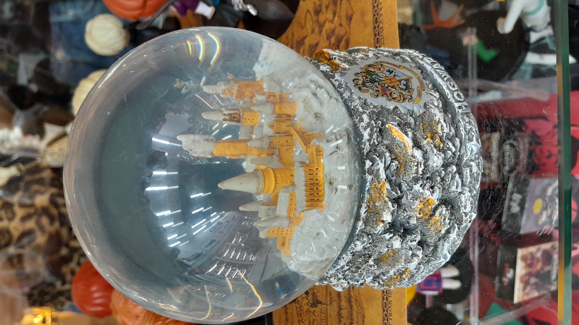 A snow globe depicting Hogwarts Castle from the Harry Potter series sits on a glass surface. The globe contains a miniature model of the castle, with snow accumulating on its spires and walls. The base of the globe is textured to resemble stone and is adorned with the Hogwarts crest and the word "HOGWARTS." Behind the snow globe, various items are visible, including a leopard-print fabric, an orange decorative item, and other indistinct objects on shelves. The overall scene suggests a retail environment, possibly a gift shop or souvenir store. This item is located in Pampatar, Venezuela.