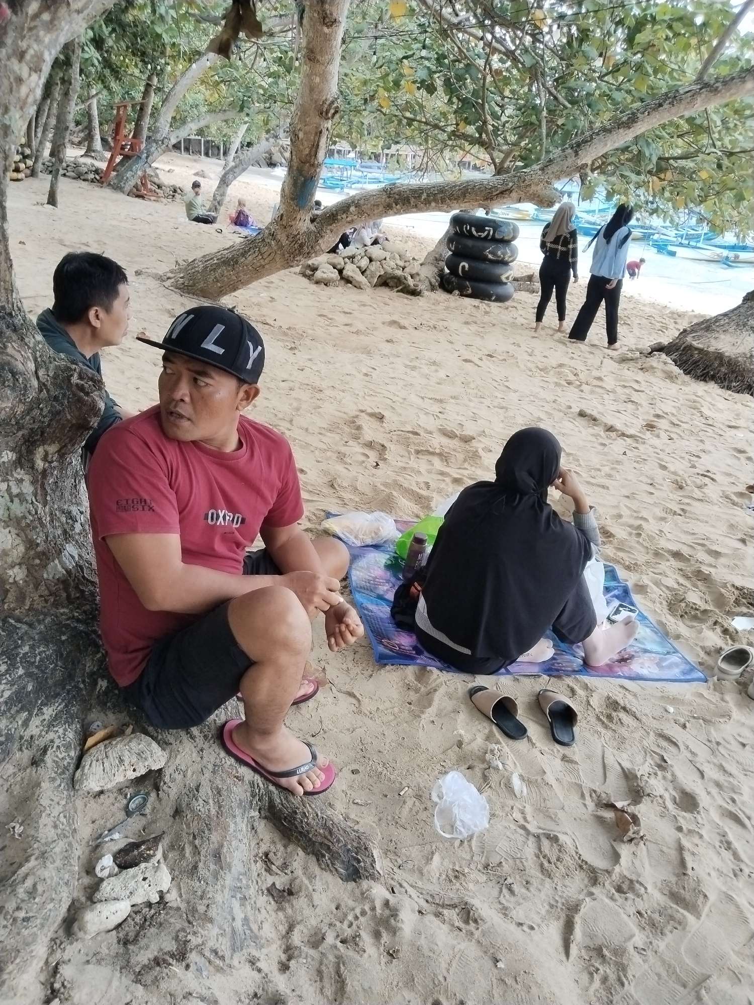 The image depicts a beach scene in Prigi, Indonesia, with several individuals present. In the foreground, a man wearing a red t-shirt and black shorts sits by the base of a tree, facing to his right and looking away. He is wearing a black baseball cap with "ONLY" written on it in white letters and pink flip-flops. To his left, partially obscured by the tree, is another person. Further back on the sand, a person wearing a black hijab and a dark outfit sits on a colorful mat, facing away from the camera and looking at their phone. Nearby, a pair of brown and black sandals are placed on the sand. In the mid-ground, a stack of black inner tubes rests near the tree line. Two individuals, both wearing headscarves, walk away from the camera towards the water. The background shows the ocean with several small boats moored. The environment is sandy, with scattered rocks and driftwood, and abundant trees providing shade over the beach.