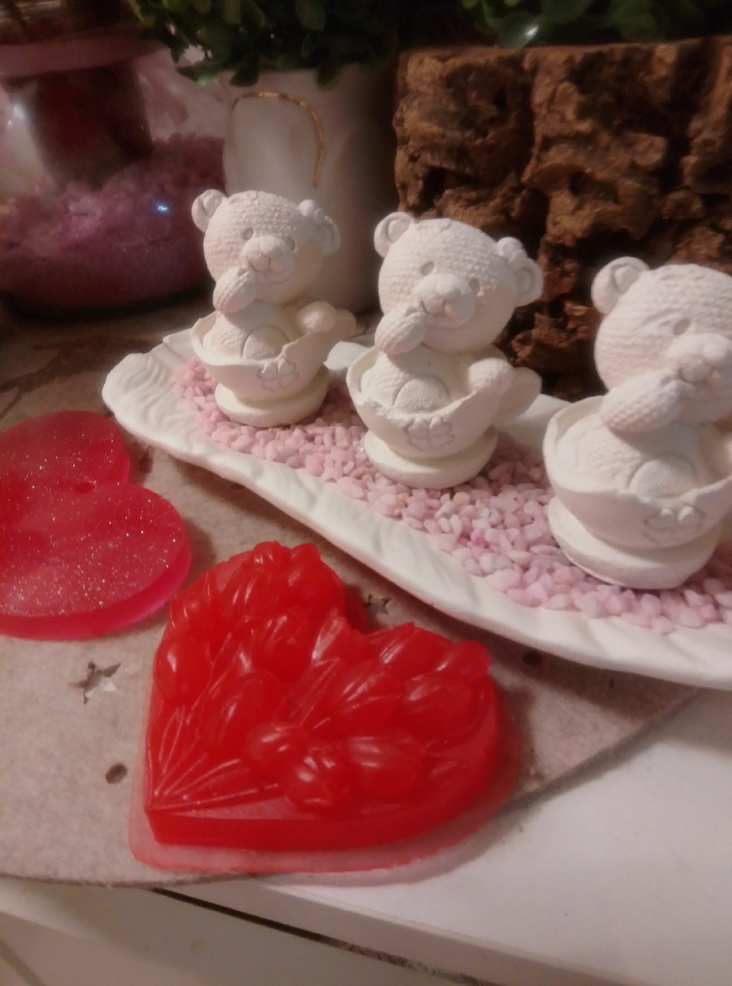 The image displays a tabletop arrangement featuring three small, white, ceramic teddy bear figurines on a white, decorative platter filled with small pink pebbles. In front of the platter, on a textured beige surface, are two red, heart-shaped objects. One appears to be a translucent soap with glitter, while the other is a more opaque, detailed soap or candy. The background shows a blurred glass jar filled with pink material, a white vase with a gold trim, green foliage, and a textured brown surface resembling bark. The scene appears to be a decorative display, possibly in a home or a retail setting. The image does not contain any information that would allow for its localization to Wedel, Germany, or any other specific city.