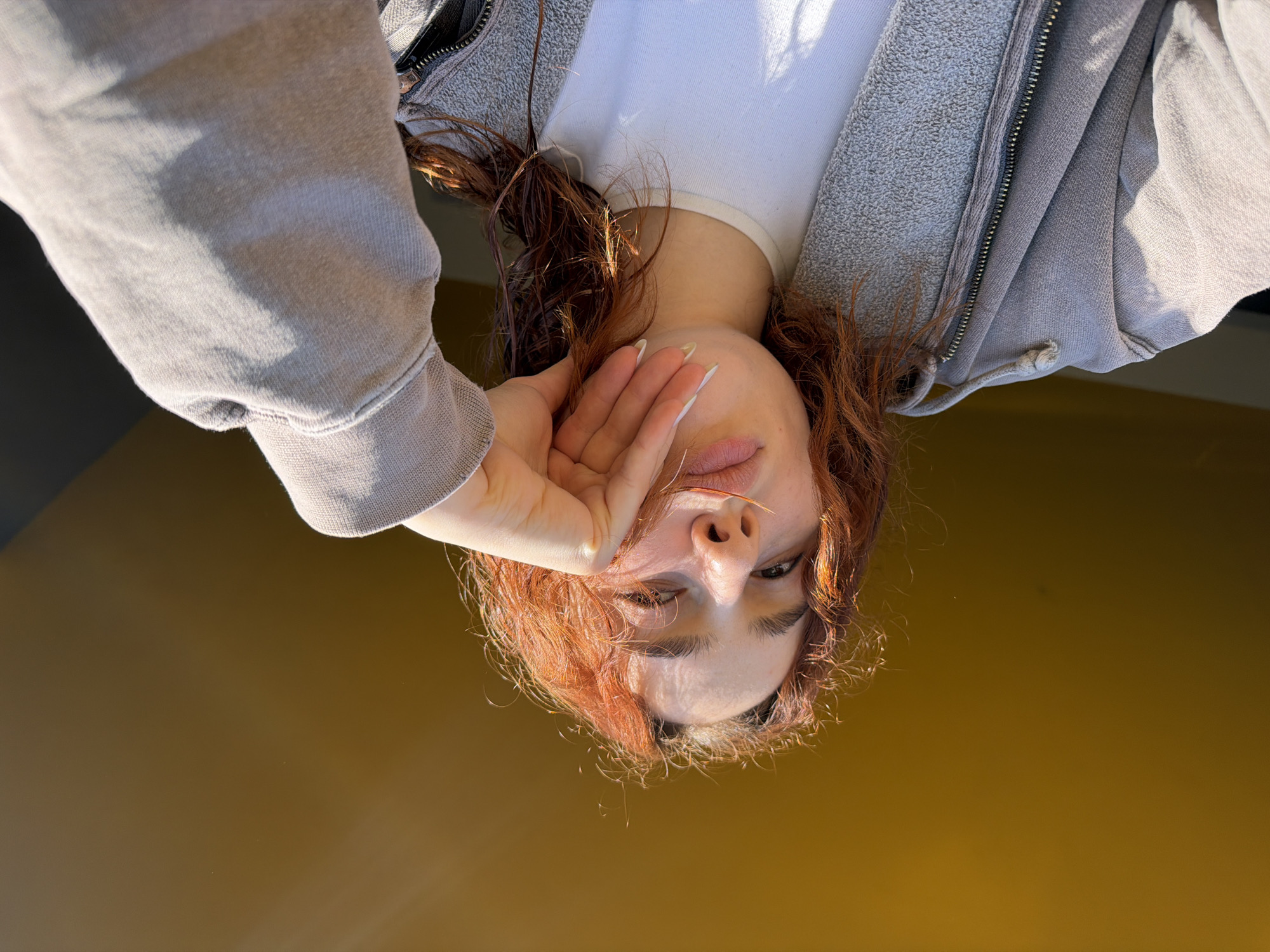 An upside-down, eye-level shot depicts a woman with reddish-brown, curly hair, fair skin, and long fingernails wearing a white t-shirt and gray zippered sweatshirt. Her hand is raised to her face, partially obscuring it. The background is a solid, dark-yellow color, suggesting a wall or other flat surface. No indication of Strasbourg, France.