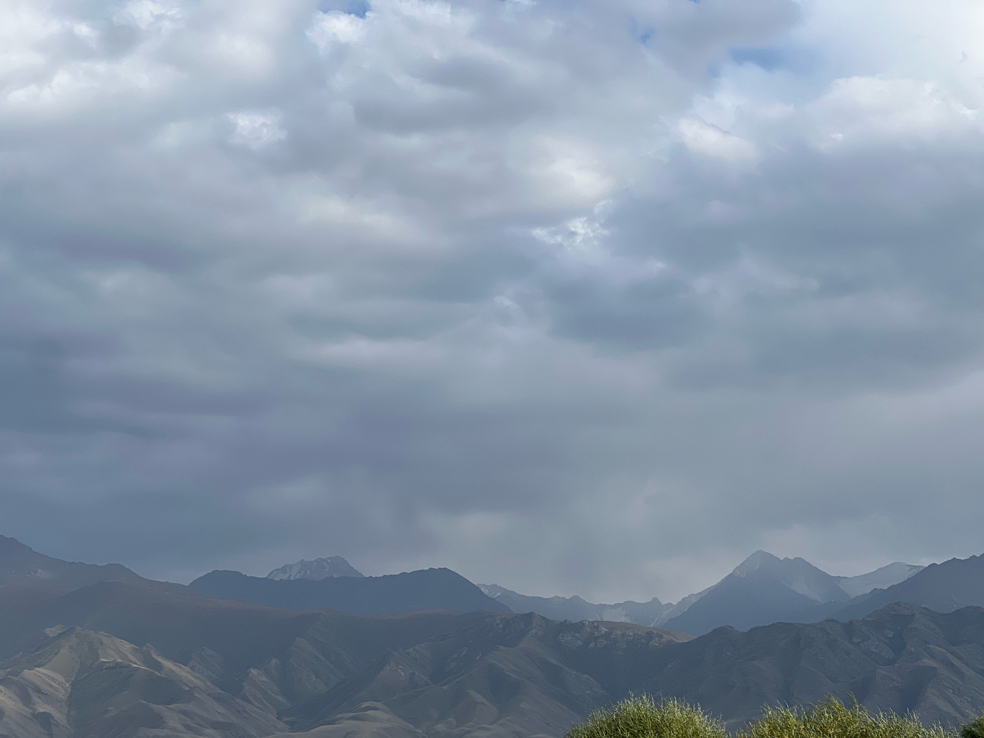 The image depicts a wide shot of mountainous terrain under a cloudy sky. The foreground is dominated by the hazy, layered peaks of mountains, with some areas showing rocky outcrops and sloped surfaces. In the far distance, the highest mountain peaks are visible, some capped with snow. The sky is filled with heavy, grey clouds, with hints of lighter blue peeking through in some areas. The overall atmosphere is subdued and overcast. There are no people, animals, or man-made structures visible in the image. The natural landscape suggests a location like Kyzyl-Tuu, Kyrgyzstan, known for its mountainous environment.