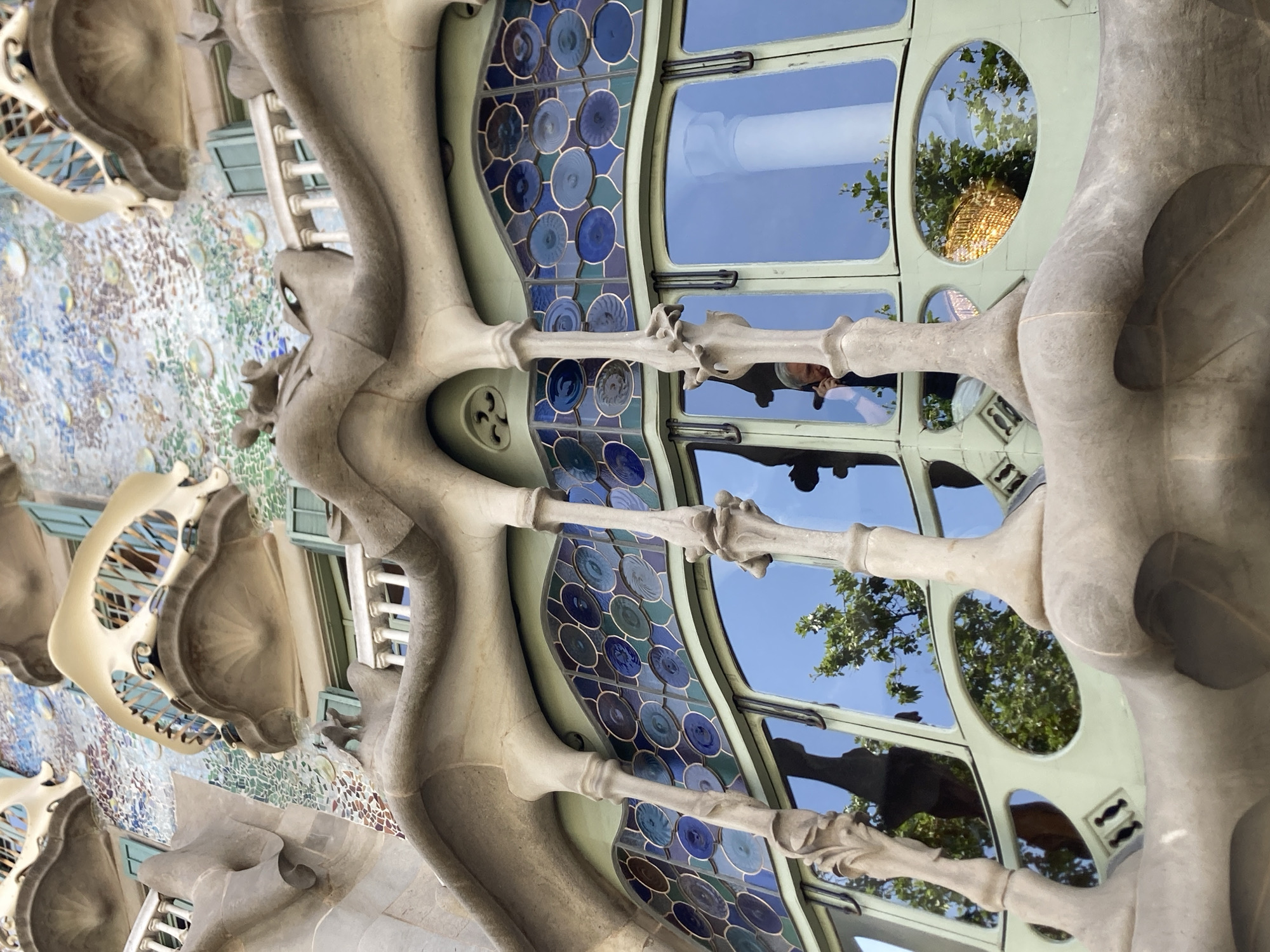 This image is a low-angle close-up shot of a detail on the exterior of Casa Batlló, a building in Barcelona, Spain, designed by Antoni Gaudí. The architecture is characterized by its organic, undulating forms and vibrant, mosaic-like tilework. The foreground features a section of a balcony with bone-like columns supporting a wavy, stone-like overhang. Behind this, there is a band of stained glass windows with circular motifs in various shades of blue and green. Large windows below the stained glass reflect the blue sky, green trees, and the facade of the building. A person is visible in the reflection of one of the large windows, appearing to hold a phone to their ear. The intricate details of the facade, including the colorful mosaic, the sculpted elements, and the unique window designs, are characteristic of Gaudí's distinctive Art Nouveau style.
