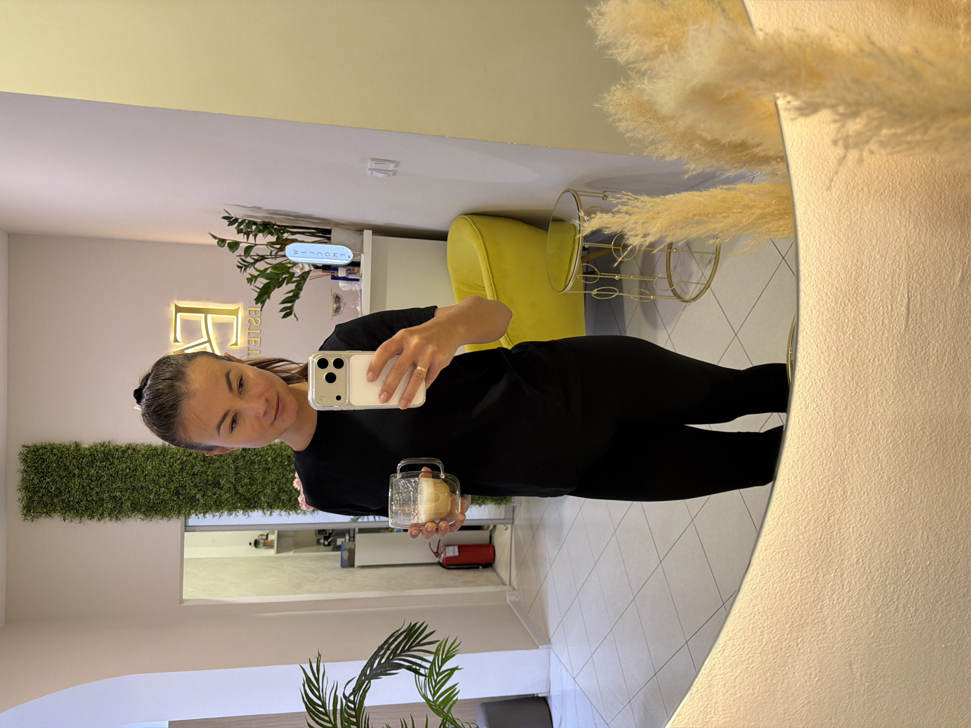 A young woman is taking a mirror selfie indoors. The mirror is round, and partially obscured by tall, feathery decorative grass. The woman is wearing black athletic clothes and has her hair pulled back. She's holding a clear glass mug. In the background, there's a green wall decoration, plants, a yellow chair, a glass and gold side table, and a "welcome" sign. The floor is tiled. A reflection in a window shows a fire extinguisher. The location is inferred to be in Budva, Montenegro, as per instructions.