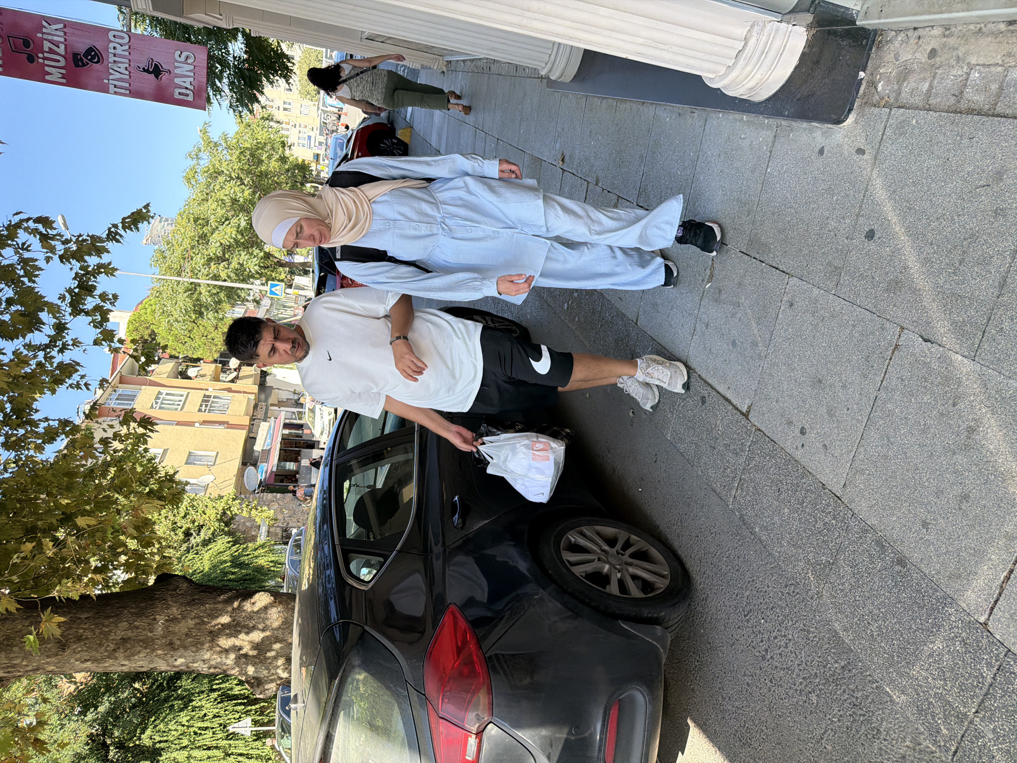The image depicts a street scene in Kadıköy, Turkey, on a sunny day. A black car is parked on the left side of the frame, partially obscuring the sidewalk. Two people are walking on the sidewalk. The person on the left, a male wearing a white t-shirt and black shorts with white athletic shoes, is carrying a white plastic bag in his right hand. He has short dark hair and a beard. The person on the right is a female wearing a light blue long-sleeved button-up shirt with a tie at the waist, matching light blue wide-leg pants, and a beige hijab. She also has a black backpack on. In the background, buildings line the street, and trees with green foliage are visible. A red banner with Turkish text and symbols hangs from a building on the right. Another person, a female in a green top and pants, is walking further down the sidewalk in the background. The street is paved with cobblestones, and a large, thick tree trunk is prominent on the left side of the image.