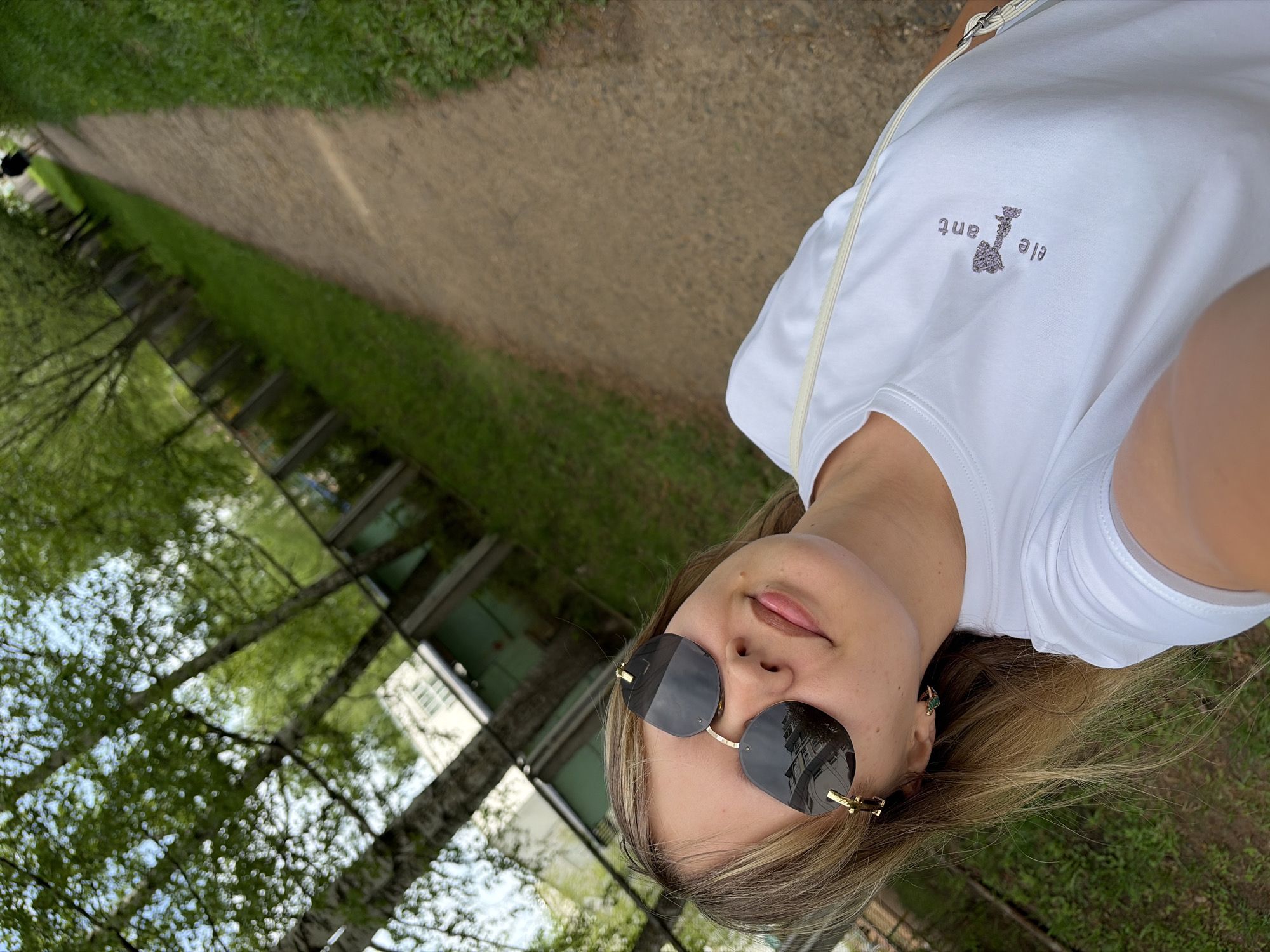 A selfie shows a woman in sunglasses, a white t-shirt, and a shoulder bag outside. The location is Chaikovsky, Russia. She is standing next to a walkway and a building with trees reflected in its window panels.