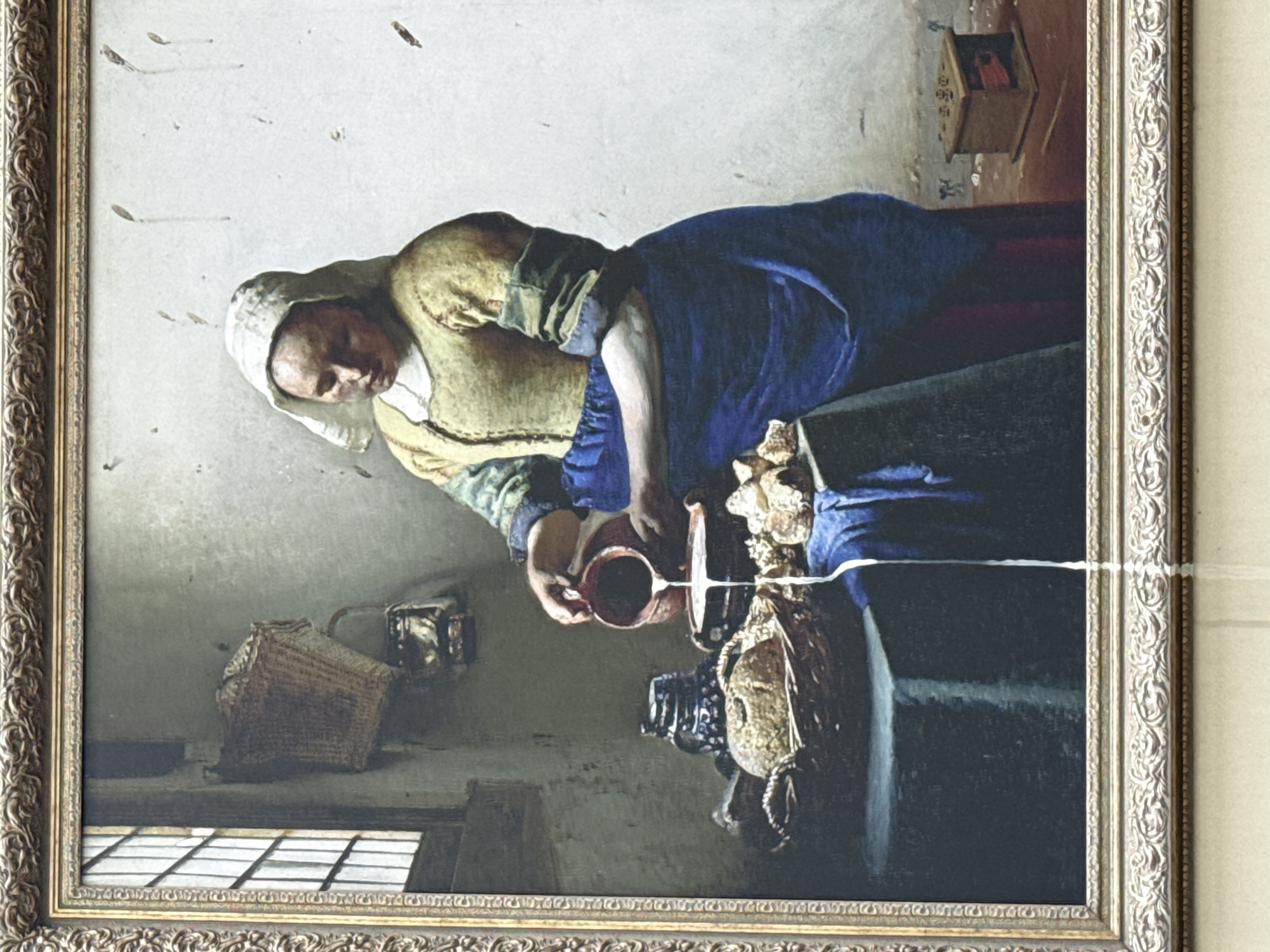 The image shows a framed painting, likely in a gallery or museum. The painting itself depicts a woman pouring milk from a terracotta jug into a bowl. The style of the painting suggests it is a work by Johannes Vermeer. The woman wears a white head covering, a yellow jacket, and a blue dress. In the background, a basket and other objects are visible. A birdhouse hangs on the wall behind her. The lower left corner of the painting shows a window. There is a thin white line that appears to be reflective or a minor defect on the painting's surface. Given the provenance of Vermeer's artwork in European collections, it is unlikely to be located in Moscow, Russia.