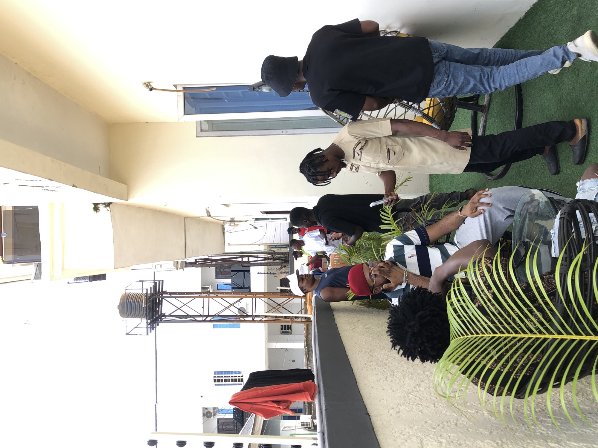 This image captures an outdoor gathering of several men on what appears to be a rooftop or balcony area during a bright day. The image is taken in portrait orientation but displayed rotated 90 degrees counter-clockwise.
In the immediate foreground, a vibrant green palm frond is prominent on the right. To its left, a man with dark, curly hair is partially visible. Next to him, a man wearing a striped polo shirt (green, white, and navy), grey sweatpants, and a red cap is seated, resting his head in his right hand. He is positioned behind a glass surface, possibly a table.
In the mid-ground, a man in a black bucket hat, black t-shirt, and blue jeans is standing, leaning slightly on a metal structure that resembles part of a bicycle or a circular stand. In front of him, another man with braided hair, dressed in a beige short-sleeved collared shirt and black trousers, also stands. The ground in this area is covered with green, artificial turf. Further into the mid-ground, more individuals are seen socializing; one man is wearing a red t-shirt, and another a white t-shirt. To the left, black and red fabrics are draped over a railing.
The background reveals a bustling urban or semi-urban environment with multiple buildings, some featuring windows and air conditioning units. A tall metal structure, likely a water tower or cell tower, stands out against the sky. The bright lighting suggests it is daytime, and shadows indicate clear weather. The overall mood appears relaxed and social, with people gathered in conversation or leisure. No visible text is discernible in the image.