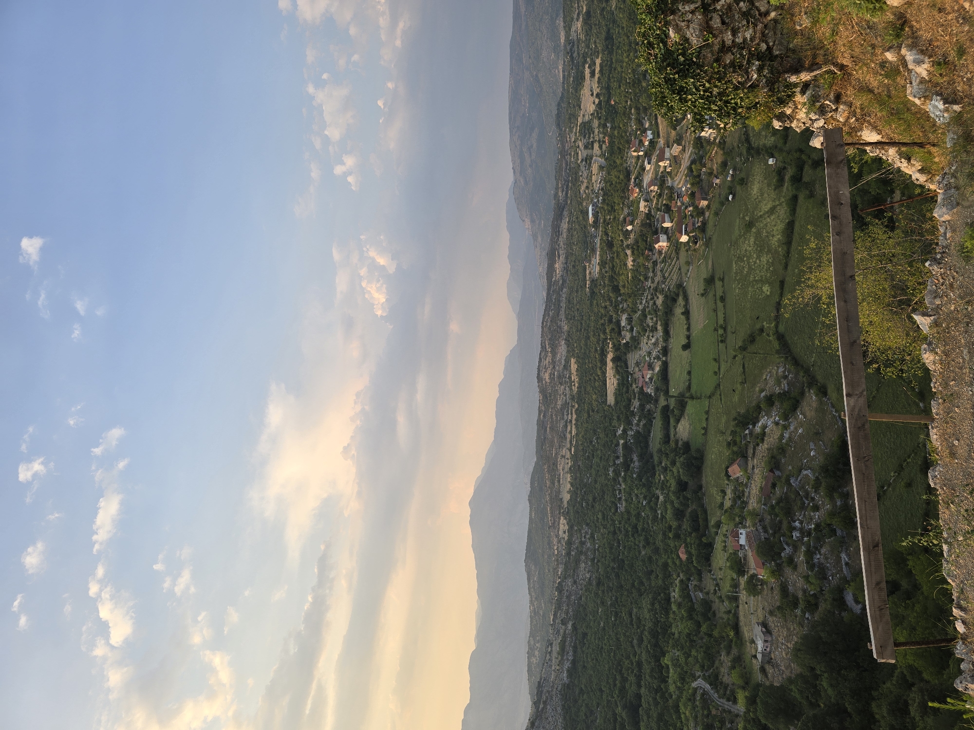 The image depicts a panoramic view of a rural landscape, likely the area surrounding Medun, Montenegro, during the late afternoon or early evening. The sky is a blend of pale blue and soft, warm hues from the setting sun, with scattered white clouds. In the foreground, a rustic wooden fence is visible, constructed from rough-hewn planks and supported by metal posts. Beyond the fence, the terrain slopes downwards, revealing a patchwork of green fields, dense foliage, and scattered houses with red-tiled roofs forming a small village. The houses are of varying sizes and architectural styles, nestled amongst trees and on small hills. In the mid-ground, the landscape is characterized by rolling hills covered in a mix of trees and shrubs, with some rocky outcrops. Distant mountain ranges, appearing hazy and muted in the fading light, form the backdrop of the scene. The overall atmosphere is serene and tranquil, with a sense of vastness and natural beauty. There are no people visible in the image.