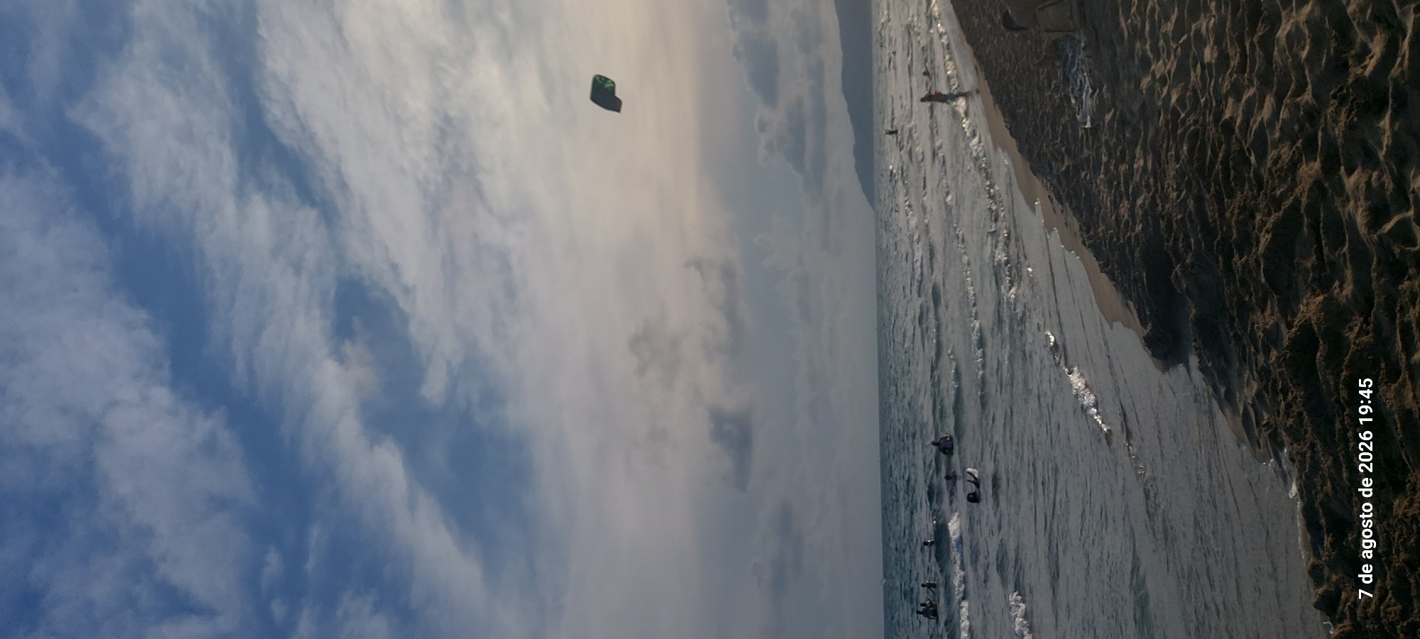 This is a photograph taken from the beach in Castelldefels, Spain, at 19:45 on August 7, 2026. The image captures a wide view of the Mediterranean Sea and the shore under a cloudy sky. The sea is calm with small waves rolling onto the sandy beach. Several people are visible in the water, some swimming and others wading. A person is seen on the shore in the mid-ground to the right. In the upper right portion of the sky, a kite, appearing dark with green accents, is flying. The foreground is dominated by the textured sand of the beach. The sun is low in the sky, casting a soft, diffused light.