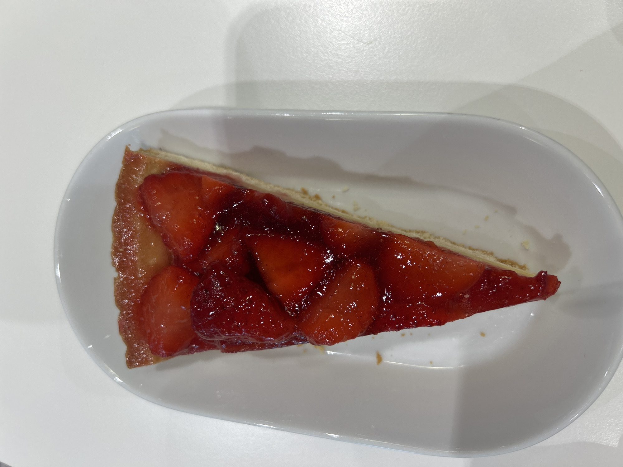 A slice of strawberry tart is presented on an oblong white plate on a white table. The tart has a golden crust, a layer of pastry cream or a similar base, and a topping of sliced strawberries glazed with a transparent jelly or glaze. The location is not discernible from the image.