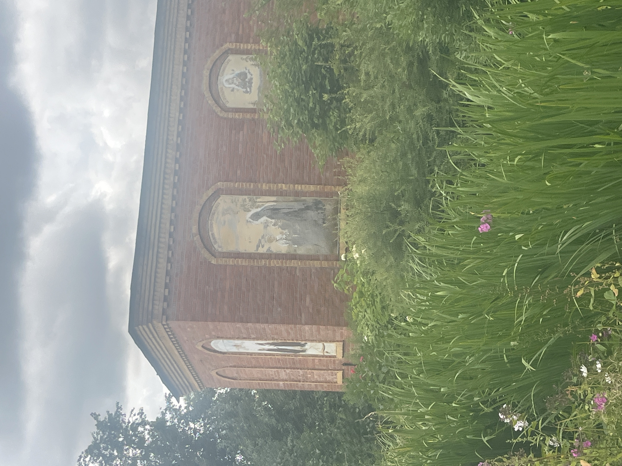 The image depicts the exterior of a brick building, likely part of a church or chapel, in Peterhof, Russia. The building has arched windows or niches containing what appear to be religious murals. The foreground is filled with lush green foliage, including tall grasses and flowering plants. The sky is overcast with dramatic clouds. The building itself is made of red brick with a decorative cornice. One mural shows a cloaked figure, and another depicts a seated figure with a halo. The overall scene suggests a somewhat overgrown, historical site.