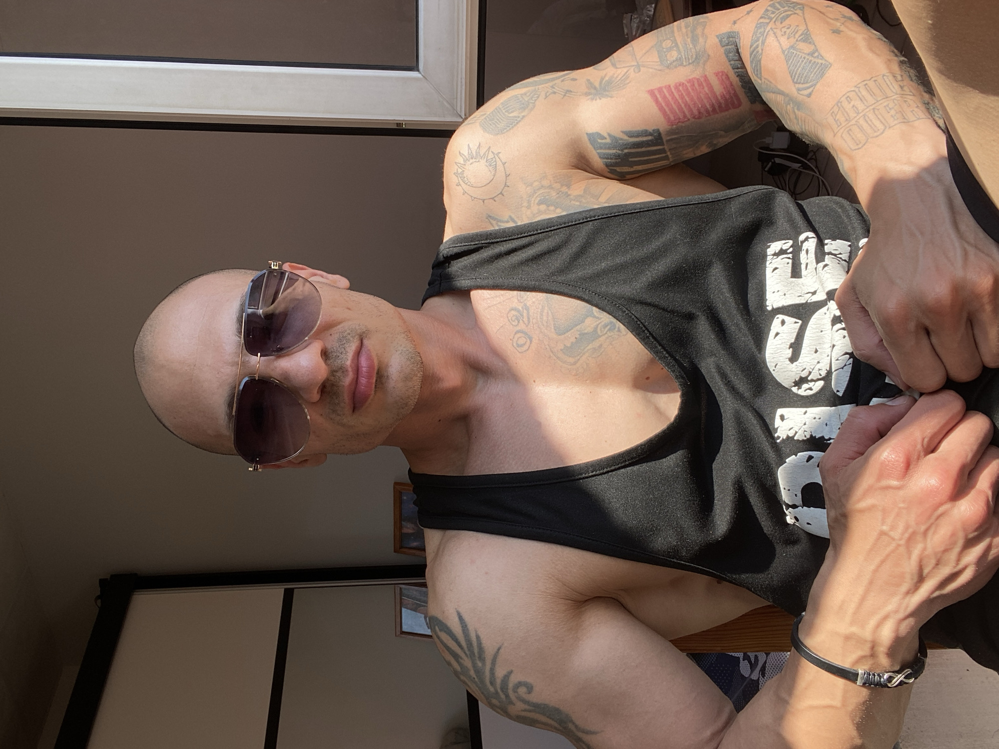 A man with a shaved head and sunglasses poses for a photograph indoors. He is wearing a black tank top that reveals tattoos on his chest, arms, and shoulders. A black bracelet is visible on his left wrist. The background shows a window frame and a glimpse of interior elements such as walls and furniture. The photo's setting is unspecified within the city of Pleven, Bulgaria.