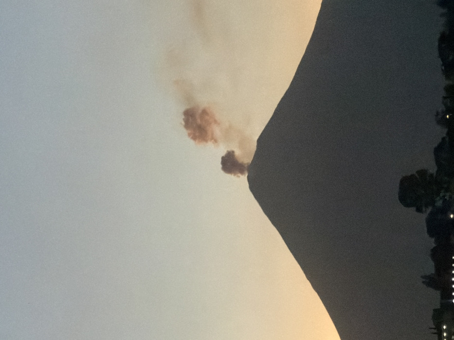 A volcano erupts, emitting a cloud of smoke. The volcano's dark silhouette contrasts with the light gray sky. Some trees and lights can be seen in the right corner of the image.