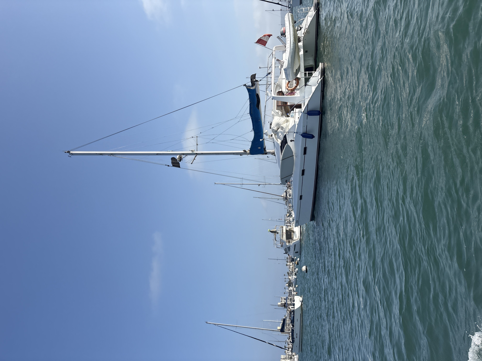 The image shows a marina in La Punta, Peru on a sunny day. The focus is on a white sailboat moored in the water. The boat has a blue sail furled at the mast, a red flag, and appears to have at least one person on board. Numerous other sailboats are visible in the background, lined up along the harbor. The water is a greenish-blue color with ripples across the surface. The sky is a clear blue with some sparse clouds.