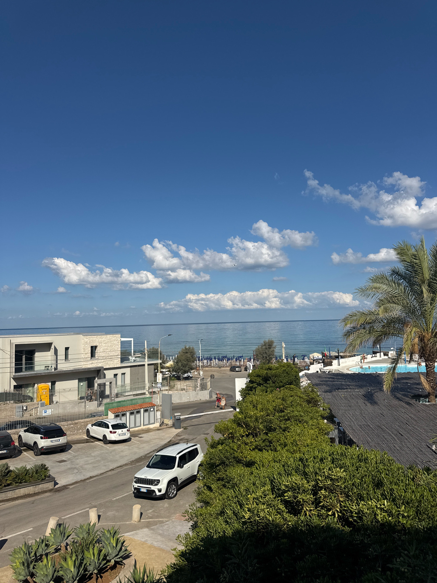 The image captures a sunny day in Capaci, Italy, overlooking the Mediterranean Sea. In the foreground, lush green trees and shrubs line a paved area with several parked cars, including a white Jeep Renegade. Behind the trees, a white building with a modern design is visible, featuring large windows and a balcony. Further in the background, the azure sea stretches to the horizon, meeting a bright blue sky dotted with white, fluffy clouds. Beach umbrellas and loungers are set up on the sandy shoreline, suggesting a resort or beachside establishment. A swimming pool with people enjoying a dip is also visible, along with a palm tree providing shade. The scene appears to be a vacation or leisure destination, with clear weather conducive to outdoor activities.
