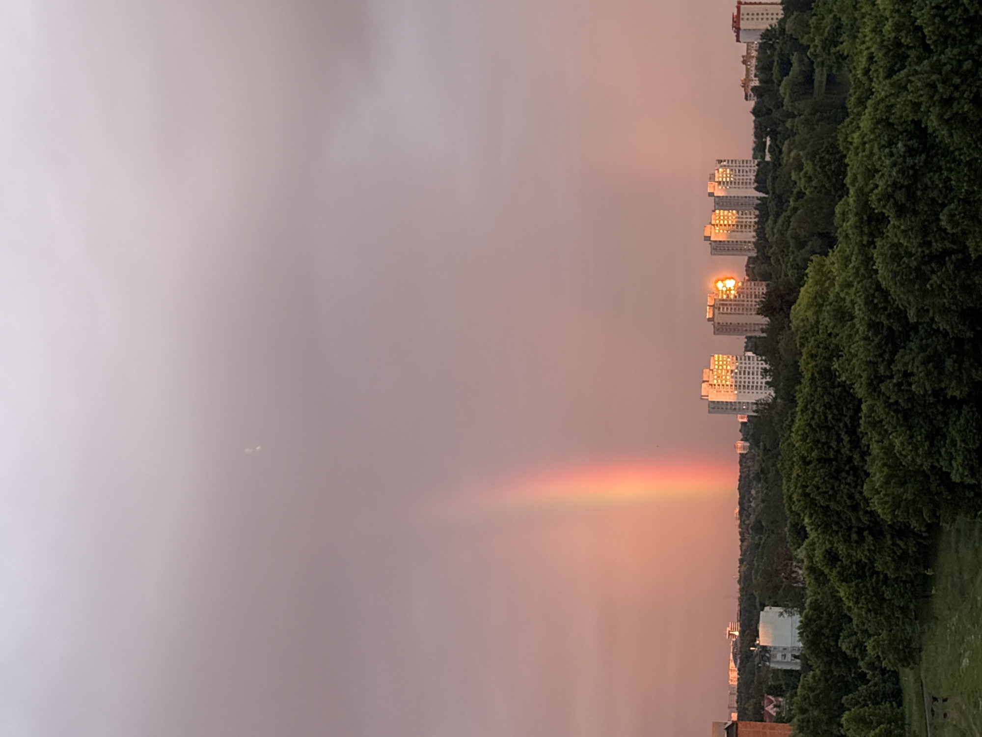 This image depicts an urban landscape in Minsk, Belarus, during what appears to be late afternoon or early evening. The sky transitions vertically from a muted purplish-gray at the top to a warm, peachy-pink hue near the horizon. A prominent, vibrant primary rainbow arcs across the left-center of the sky, displaying distinct spectral colors. A fainter, secondary rainbow is subtly visible to the right of the primary one, blending with the pink sky.
The lower half and midground are dominated by a dense, dark green canopy of deciduous trees. Beyond this foliage, a cluster of multi-story buildings is visible on the right, with their windows intensely reflecting golden sunlight, creating prominent solar flares, particularly on the central building within the cluster. Additional residential or commercial structures are sparsely distributed in the distant background, including a partial view of a reddish-brown building on the far left and a smaller, white building partially obscured by trees in the mid-left. No individuals, specific vehicles, or discernible actions are present.