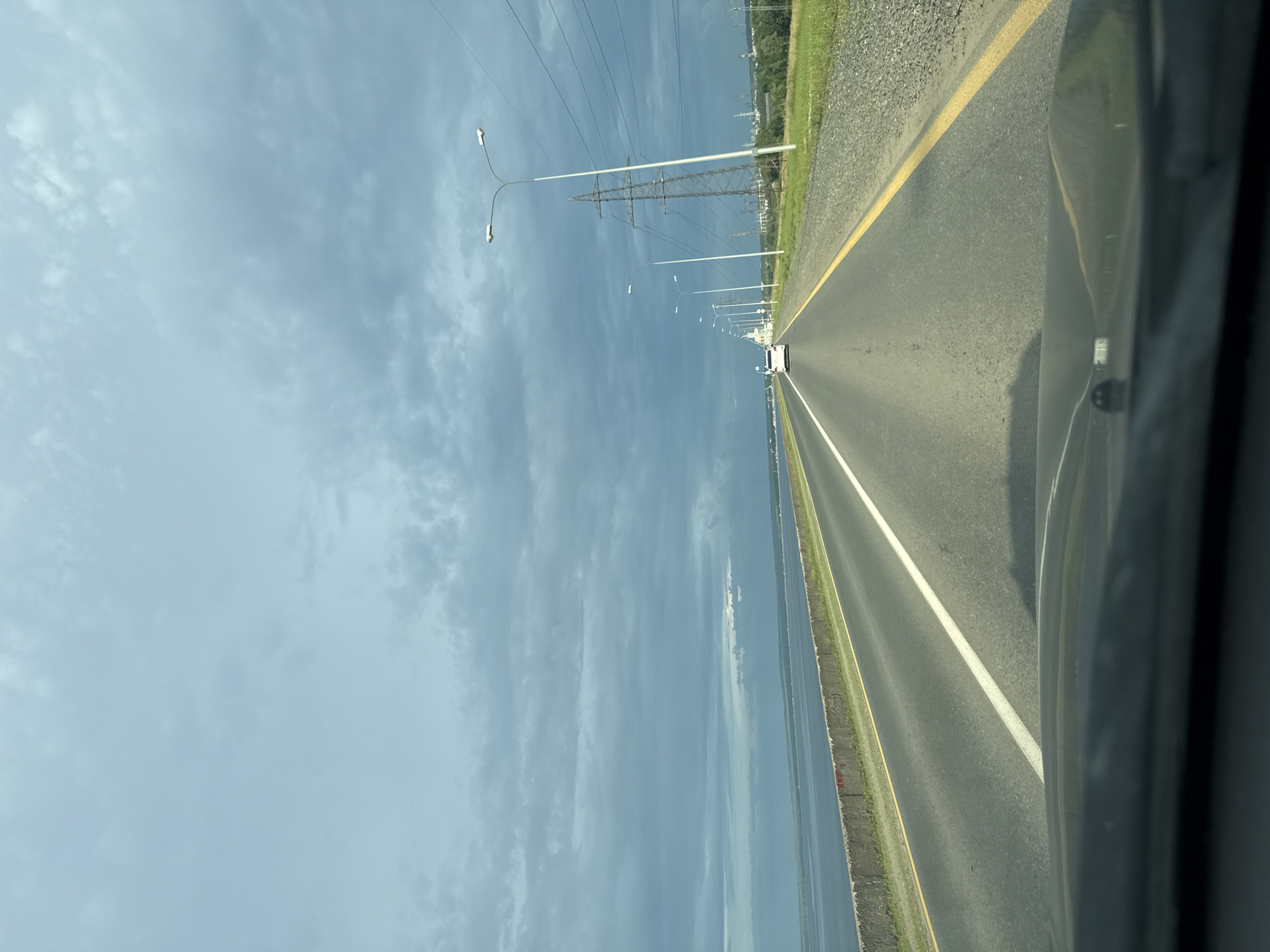 An image captured from inside a vehicle, looking out over a highway. The highway runs parallel to a large body of water, likely a river or lake, on the left side of the road. A concrete barrier separates the road from the water. On the right side of the highway, there is a grassy verge, power lines supported by tall pylons, and distant industrial structures. The sky is overcast with dark, dramatic clouds. A single white car is visible ahead on the highway. The lower portion of the image shows the dashboard and windshield of the vehicle. The location is identified as Noviy, Russia.