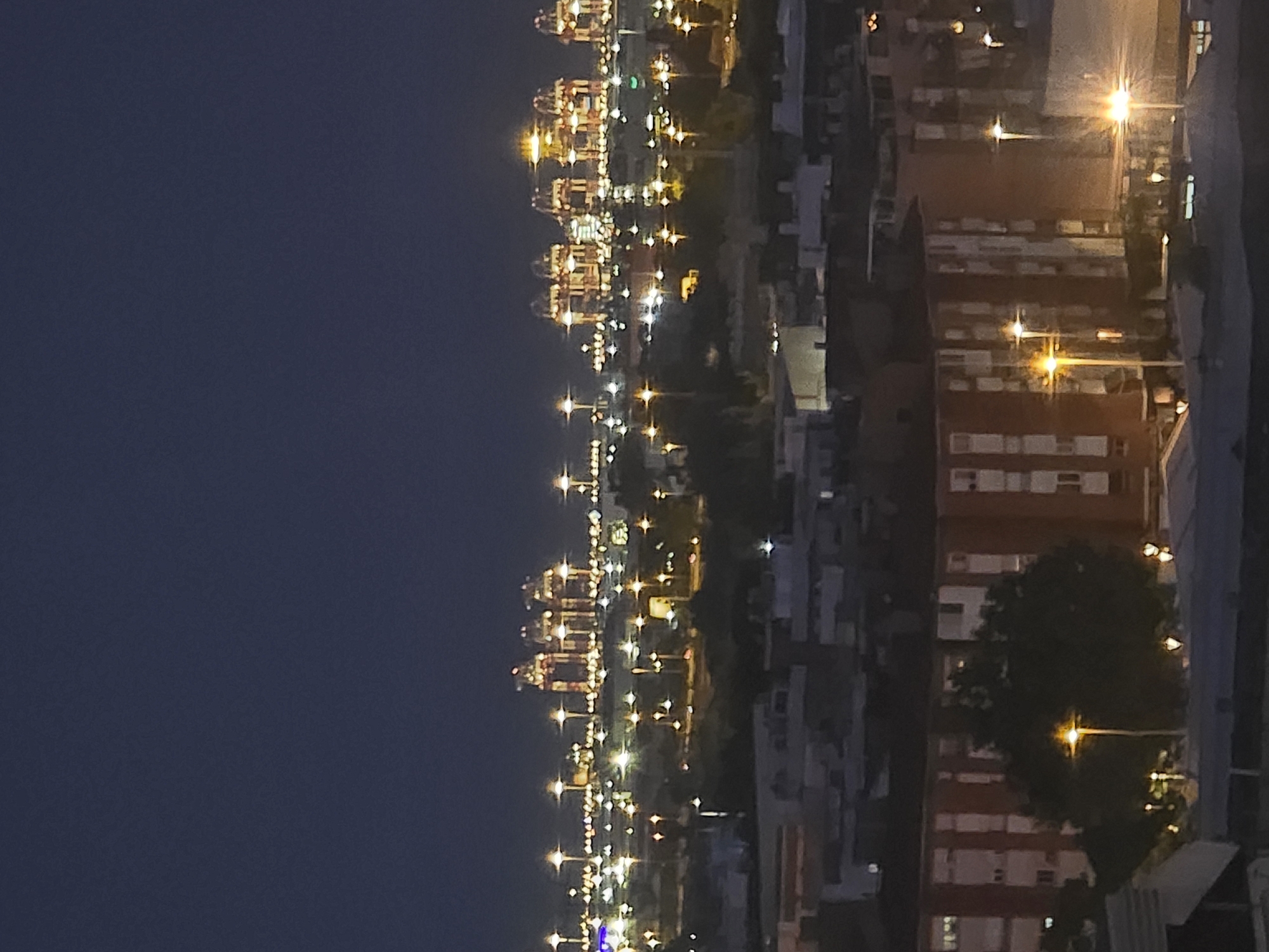 The image depicts a nighttime view of the port of Barcelona, as seen from Cornellà de Llobregat, Spain. The foreground shows the rooftops of buildings in Cornellà, with streetlights illuminating the scene. The middle ground features more residential buildings and trees. In the background, the Port of Barcelona is visible, with its prominent cranes illuminated by numerous lights. The sky is dark, indicating it is late evening or night. The overall impression is that of an urban landscape at night, with the industrial port activity creating a stark contrast to the quieter residential areas.