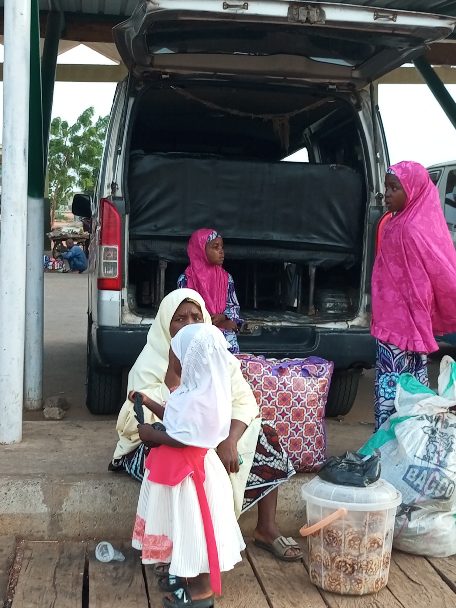 In Gombe, Nigeria, a group of people are gathered near a white van with its rear doors open. The scene appears to be outdoors, possibly a marketplace or a loading area.
Three women are prominently visible. One woman, wearing a vibrant pink hijab and patterned dress, stands to the right, facing left. Another woman, dressed in a pale yellow hijab and a patterned wrap skirt, is seated on a step, holding a black object. A young child, wearing a white dress with pink trim and a white head covering, stands in front of the seated woman, also holding a black object. A fourth person, a young girl in a pink hijab and patterned dress, is seated on the back of the van. In the background, a man in a blue outfit is hunched over a cart with various items.
Objects in the scene include the van itself, a patterned purple and orange tote bag, a clear plastic bucket filled with food items, and two large white sacks, one with the visible word "BACCO". A plastic bottle is on the ground in the lower left. The environment is characterized by a concrete walkway or step, wooden planks, and a metal roof overhead, supported by a green-and-white striped pole. Trees and foliage are visible in the background. The overall impression is of a moment of transit or transaction.