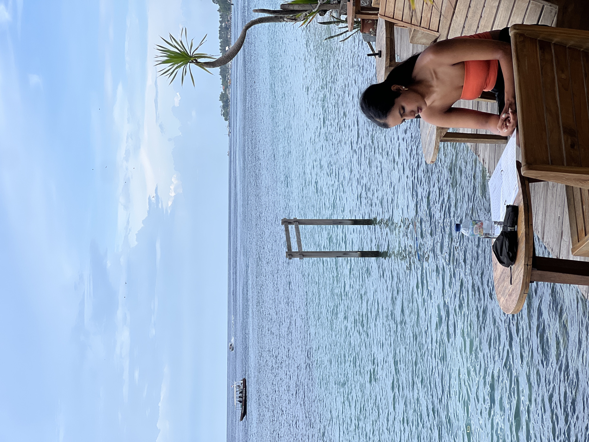 This image captures a serene waterside scene, likely in Nusa Penida, Indonesia. The primary subject is a woman with dark hair, dressed in an orange strapless top and dark pants. She is seated on a wooden chair at a round wooden table on a deck or pier built over water, leaning forward and looking down at something on the table. Her hands are clasped lightly in front of her.
On the wooden table, a plastic water bottle, a black item (possibly a hat or small bag), and a white sheet of paper (likely a menu, though no specific text is legible) are visible. The wooden deck extends over a calm body of water, which appears to be a sea or large lake, with gentle ripples on its surface.
In the water, further from the deck, two wooden posts form a simple, open rectangular structure. In the distance across the water, a boat carrying several people and a smaller vessel are visible. Beyond the water, a faint outline of land or an island can be seen. A distinctive palm-like plant with long, spiky leaves grows from the wooden structure, extending its trunk and foliage over the water.
The scene is illuminated by bright, natural daylight, suggesting it is late morning or early afternoon. The sky above is light blue with scattered clouds. The overall impression is one of relaxation and leisure in a tropical, waterfront setting.
