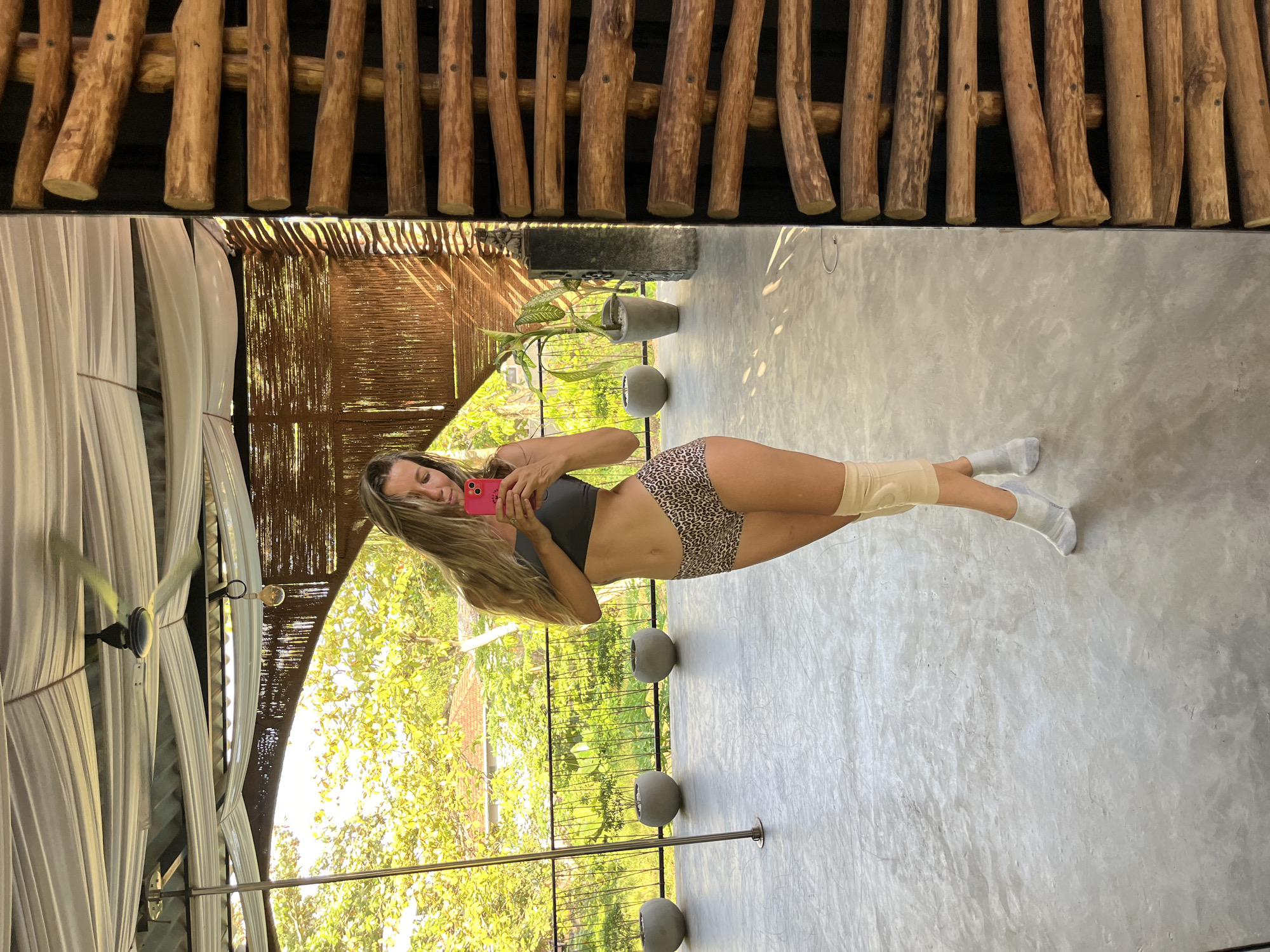 A woman is taking a selfie in a room with a concrete floor and a ceiling covered with white fabric. She is wearing a grey sports bra, leopard print shorts, a beige knee brace, and white socks. She is holding a pink smartphone in her right hand. In the background, there is a pole, potted plants, and trees visible through a railing. The room appears to be open to the outdoors. This location is likely in Weligama, Sri Lanka, given the tropical foliage and open-air architecture.
