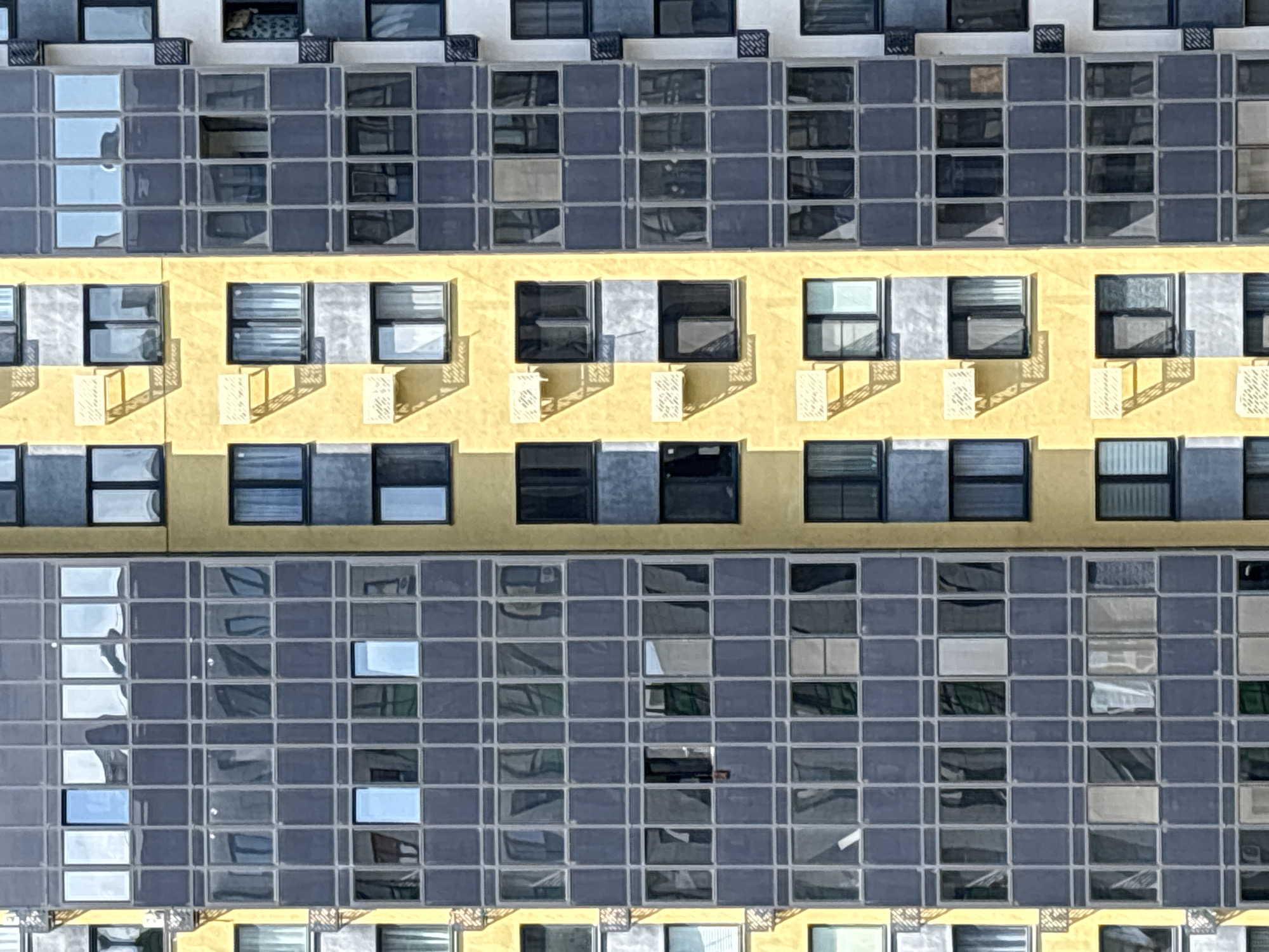 The image shows a close-up, aerial view of the facade of a modern apartment building in Kazan, Russia. The building features a repeating grid pattern of windows and balconies. The facade is divided vertically into sections of varying colors and window styles. On the left, a section of the building is clad in dark gray panels with a grid of windows. Adjacent to this, a yellow section has smaller windows and some decorative elements. Further to the right, another section features dark gray balconies and windows, and a white section with a smaller set of windows. The sunlight casts distinct shadows across the building's surfaces, indicating a clear day. No people are visible in the image.
