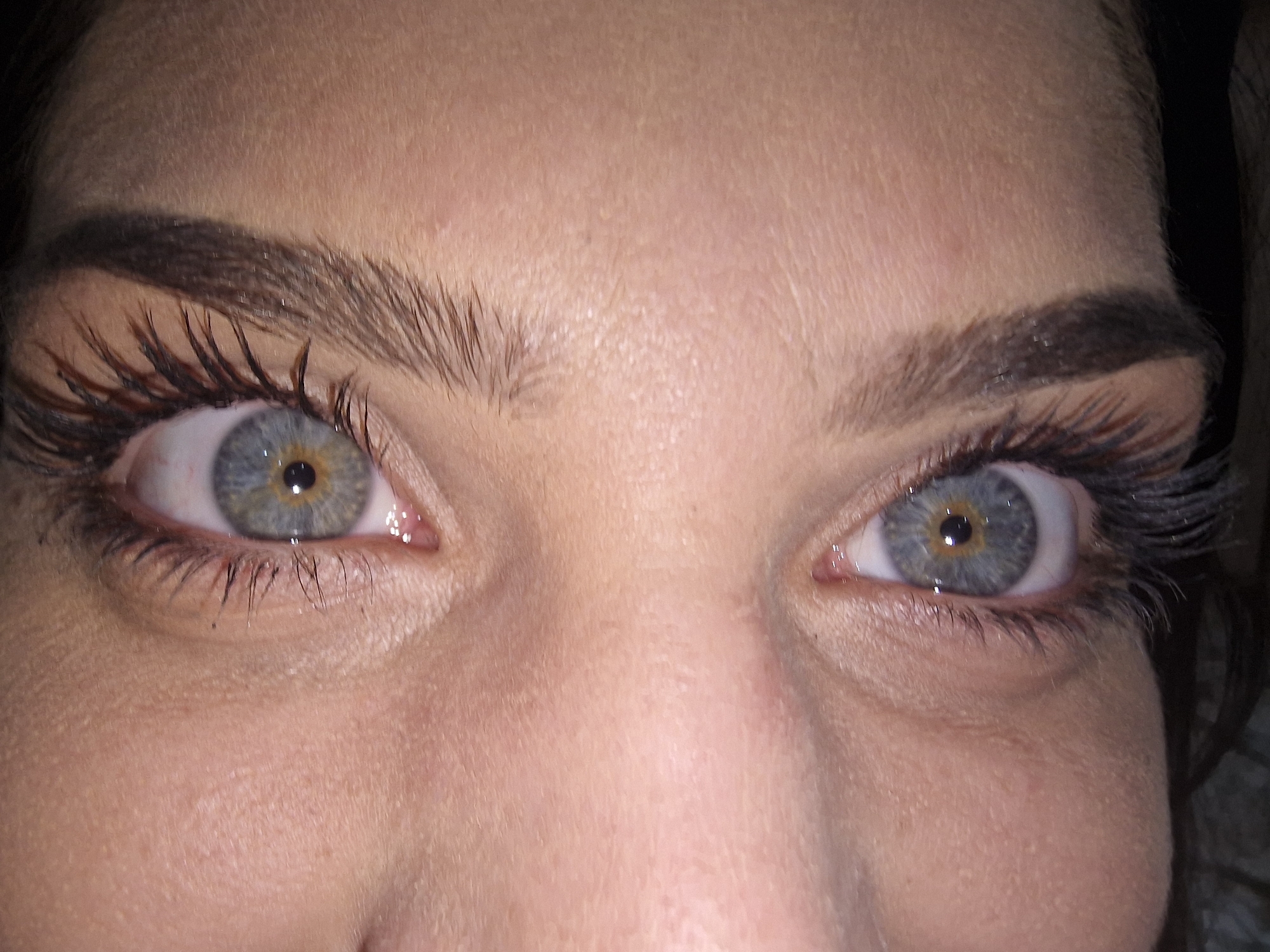 A close-up photograph depicts a woman's face, focusing on her eyes and the surrounding area. Her skin tone appears fair with visible pores and textures. She has well-defined eyebrows with individual hairs clearly visible. Her eyes are a striking grey-blue with a distinct pattern in the iris and noticeable eyelashes. The lighting in the photo is somewhat uneven. The location is unknown.
