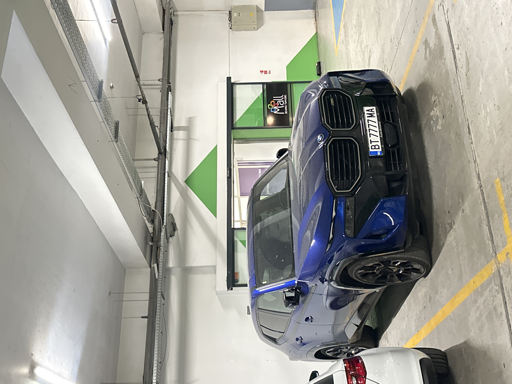 This media file captures a blue BMW parked in what appears to be an indoor parking garage or commercial area, possibly a shopping mall given the visible signage. The car is the primary subject, a modern vehicle with a sleek design, prominent grille, and a license plate reading "BT 7777 MA". The setting is functional and utilitarian, with concrete floors, exposed conduits and pipes running along the ceiling, and white walls. There are diagonal green geometric shapes painted on a section of the wall behind the car, adding a splash of color to the otherwise neutral environment. A sign for "Mall" with Bulgarian text below ("BEAMBO TUMPOBO") is visible through a glass door, suggesting a retail or entertainment complex. The lighting appears to be artificial, consistent with an indoor space. The image is taken from a low angle, emphasizing the car's presence. The overall impression is one of a contemporary commercial space housing a luxury vehicle. The orientation of the image is rotated, showing the scene from a side perspective. No people are visible, and the weather is not relevant as it is an indoor scene. The location context of Veliko Tarnovo, Bulgaria, is provided but not directly evident from visual cues within the image itself, other than the Bulgarian text on the mall sign.