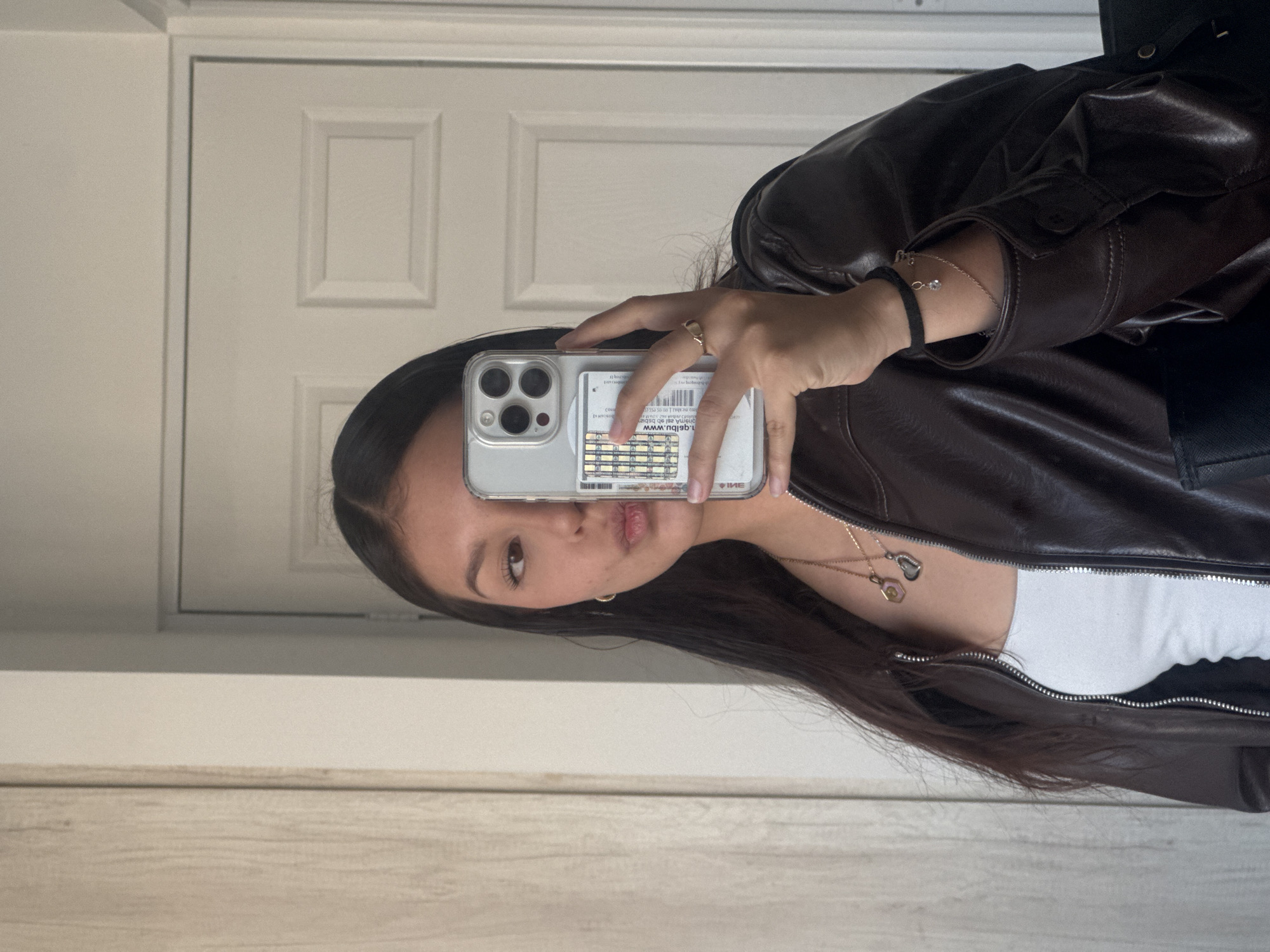 A young woman is depicted taking a mirror selfie indoors. She has long, dark hair and is wearing a dark brown faux leather jacket over a white top. Her expression is neutral, with slightly pursed lips. She holds a silver iPhone with a clear case in her right hand, revealing a gold-colored ring on her ring finger. Inside the phone case, a white card with a barcode and partially visible reversed text, including "WWW" and "IME", is visible. She wears two gold-colored necklaces with pendants, a black elastic bracelet, and a delicate gold chain bracelet with a small charm on her left wrist. The background reflects a light-colored interior door with paneling and what appears to be a light wooden floor or wainscoting. This scene is contextualized in San Francisco Ocotlán (Ocotlán), Mexico.