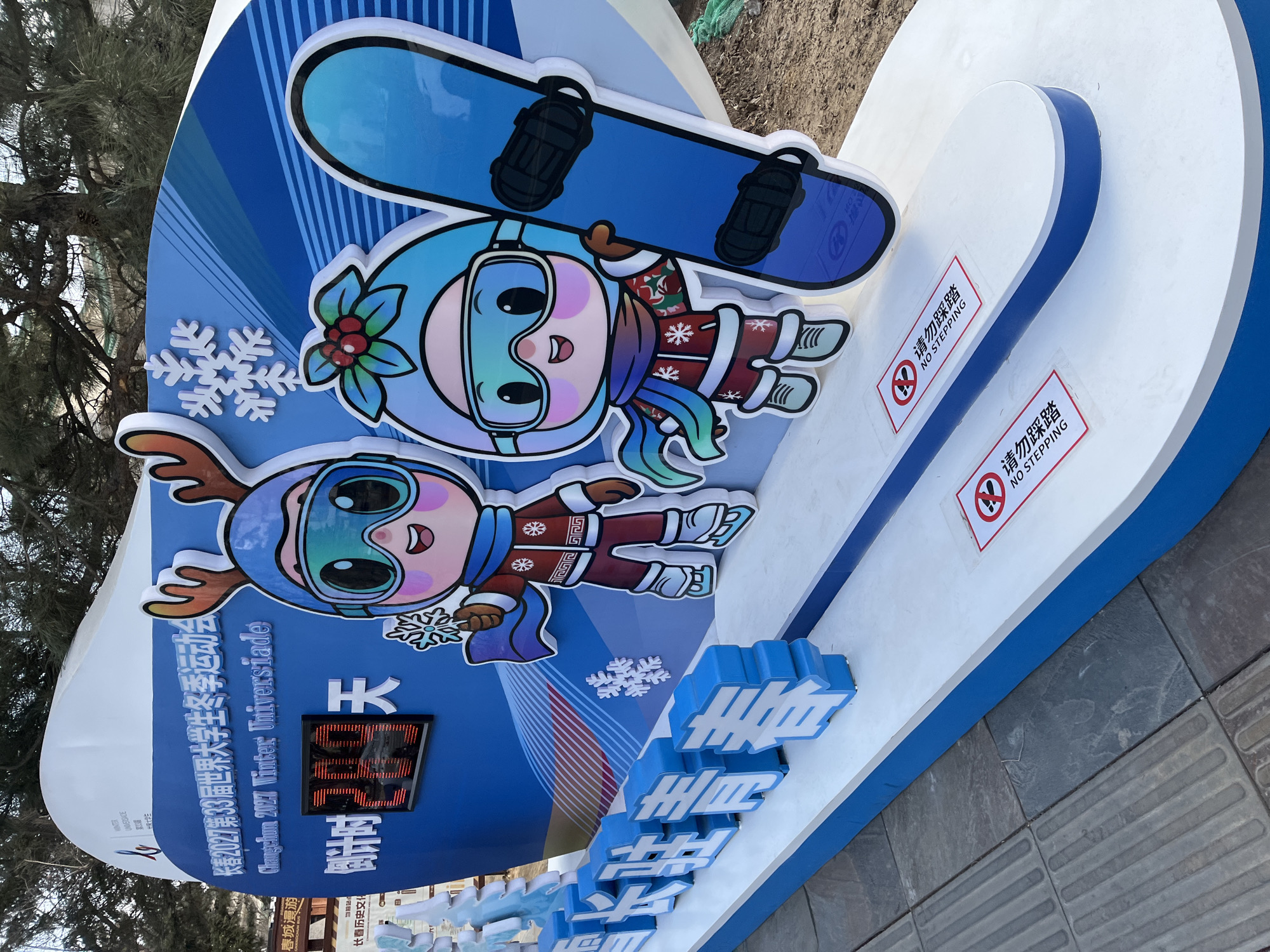 The image shows a Changchun 2027 Winter Universiade promotional sign, likely located in Changchun City, China. The sign is predominantly blue and white, featuring cartoon mascots dressed in winter gear, including goggles and winter clothing, and holding snowflakes. One mascot holds a snowboard. The sign includes Chinese text and the English translation "Changchun 2027 Winter Universiade". An electronic counter on the sign displays a number, and blue block lettering spells out text in Chinese. The sign is placed on a white platform with blue trim, and the platform has signs indicating "No Stepping" in both Chinese and English. The environment around the sign includes a paved area and foliage.