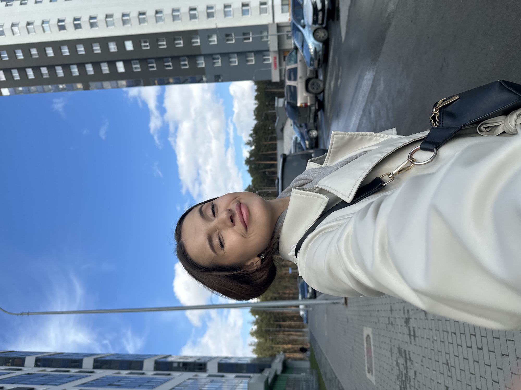A young woman with dark hair and closed eyes smiles in a selfie-style photo taken in Yekaterinburg, Russia. She is wearing a white leather jacket over a gray hooded sweatshirt and a black crossbody bag. The background shows a modern apartment building, a clear blue sky with scattered clouds, a row of pine trees, and parked cars. The ground is paved with gray bricks.