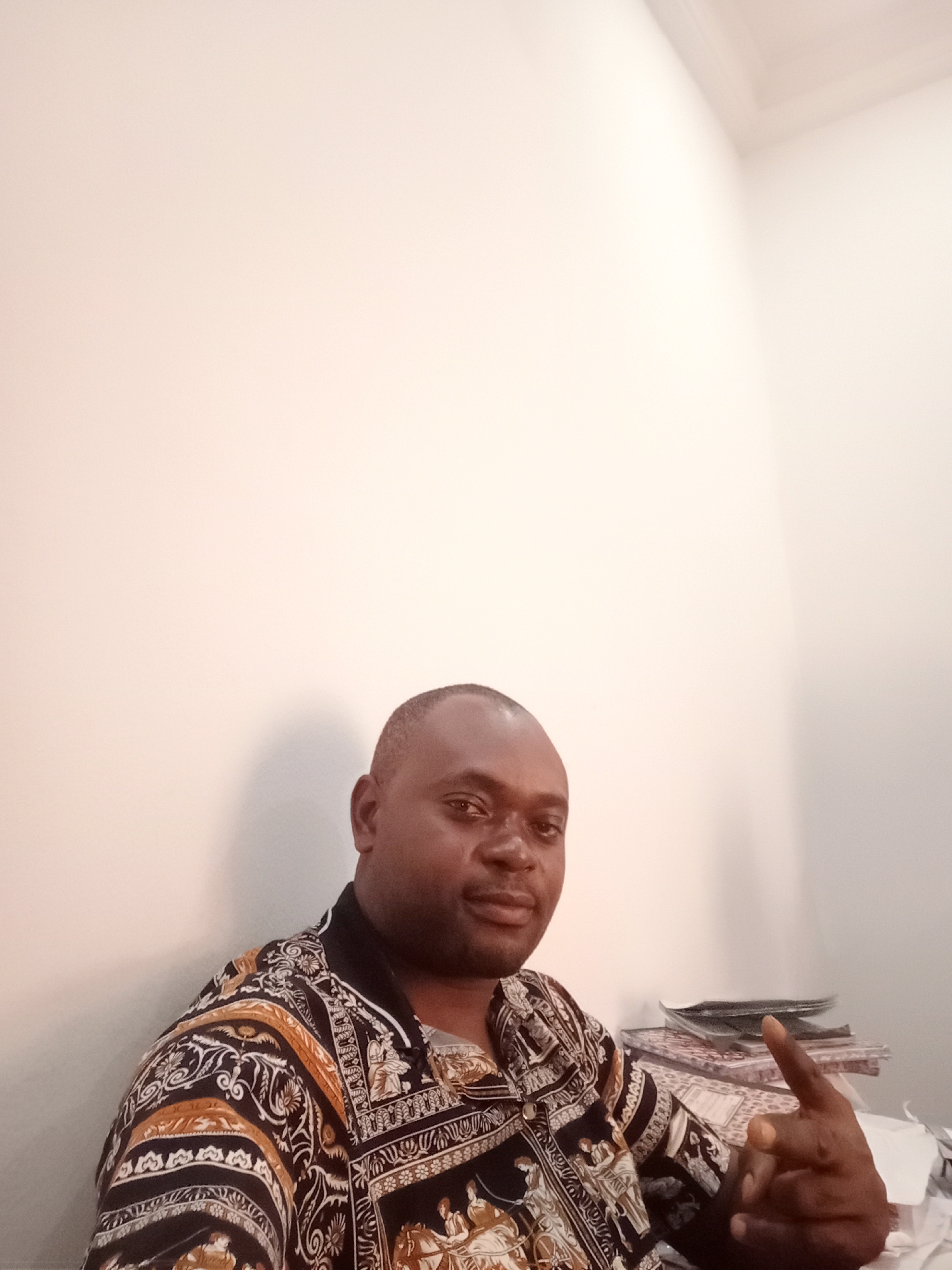 A man is seated indoors, likely in Gwarinpa, Nigeria, posing for a portrait. He is a dark-skinned individual, appearing middle-aged, with short dark hair and a neatly trimmed mustache and beard. He wears a distinctive long-sleeved collared shirt featuring an elaborate pattern in black, white, and golden-brown hues. The design includes classical or historical figures, horses, and intricate ornamental motifs.
The man looks directly at the camera with a calm, slightly smiling expression. His right hand is raised, with his index finger pointing upwards. The background is a plain, light-colored wall, indicating an indoor setting. To his right, a small stack of books, magazines, or papers rests on a surface. The lighting is soft and even, typical of an indoor environment, casting a subtle shadow behind him. No text is visible in the image.
