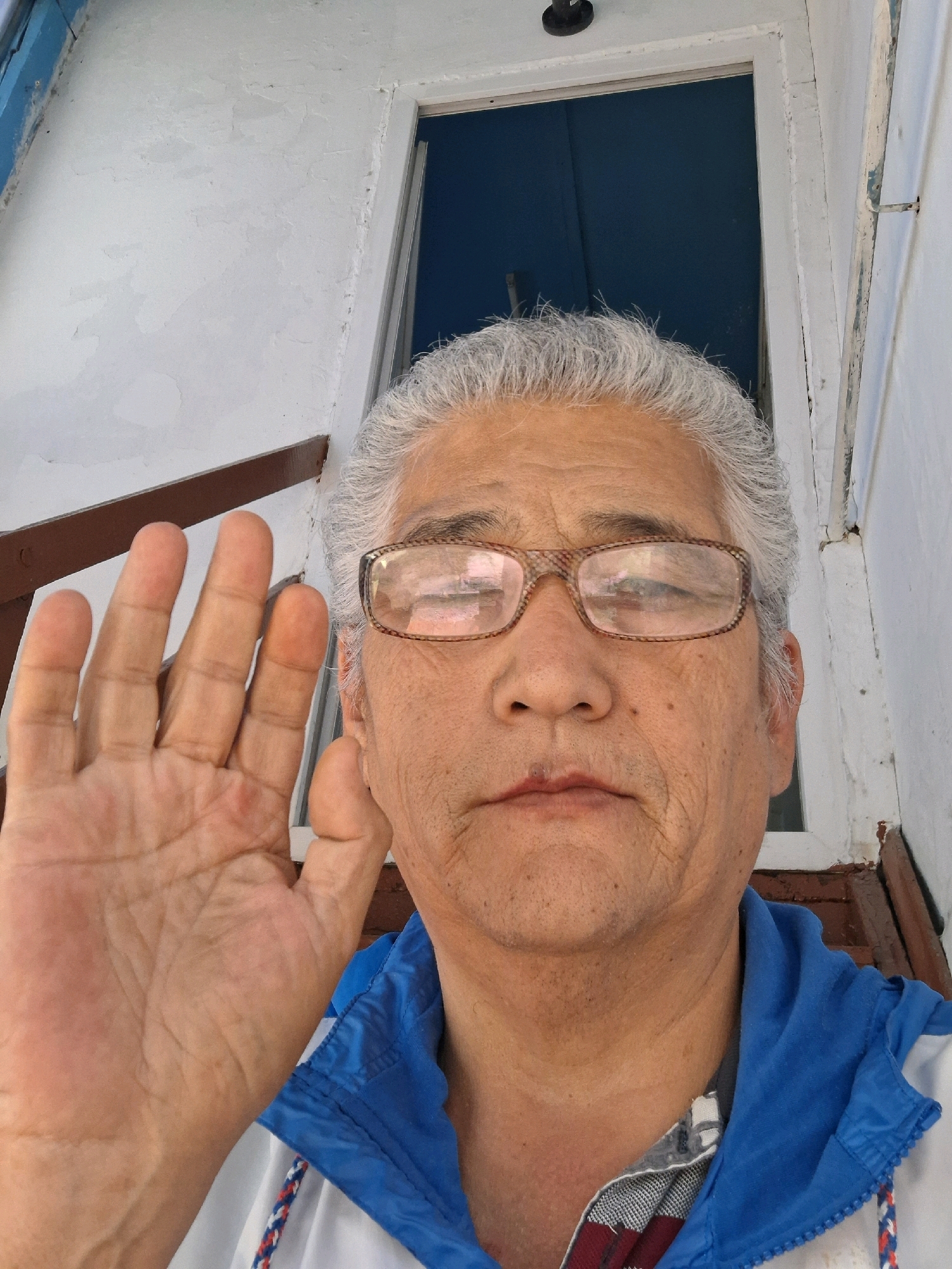 An adult male with graying hair and glasses is taking a selfie. He is wearing a blue and white jacket over a checkered shirt. His left hand is raised in front of his face, palm facing the camera. The background appears to be an exterior of a building with white walls and a dark blue door or window. There are wooden steps and a railing visible. The image does not contain enough information to determine the specific location in Almaty, Kazakhstan.