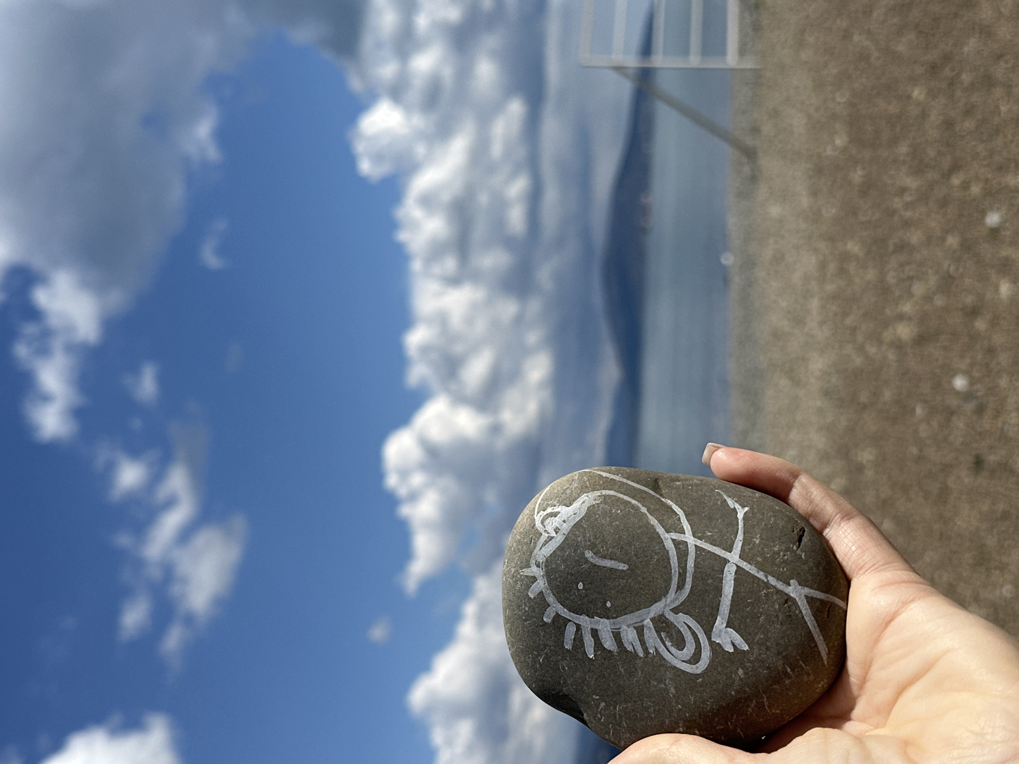 A hand holds a smooth, gray rock with a child-like drawing of a face on it. The drawing is done in white and depicts a rounded face with closed eyes, a linear mouth, and radiating lines below the face. There are also curly lines. The background features a bright blue sky with fluffy white clouds. To the right, the scene transitions into a blurry view of a beach or shoreline and a body of water. A metal structure stands on the shore. The location is Myskhako, Russia.