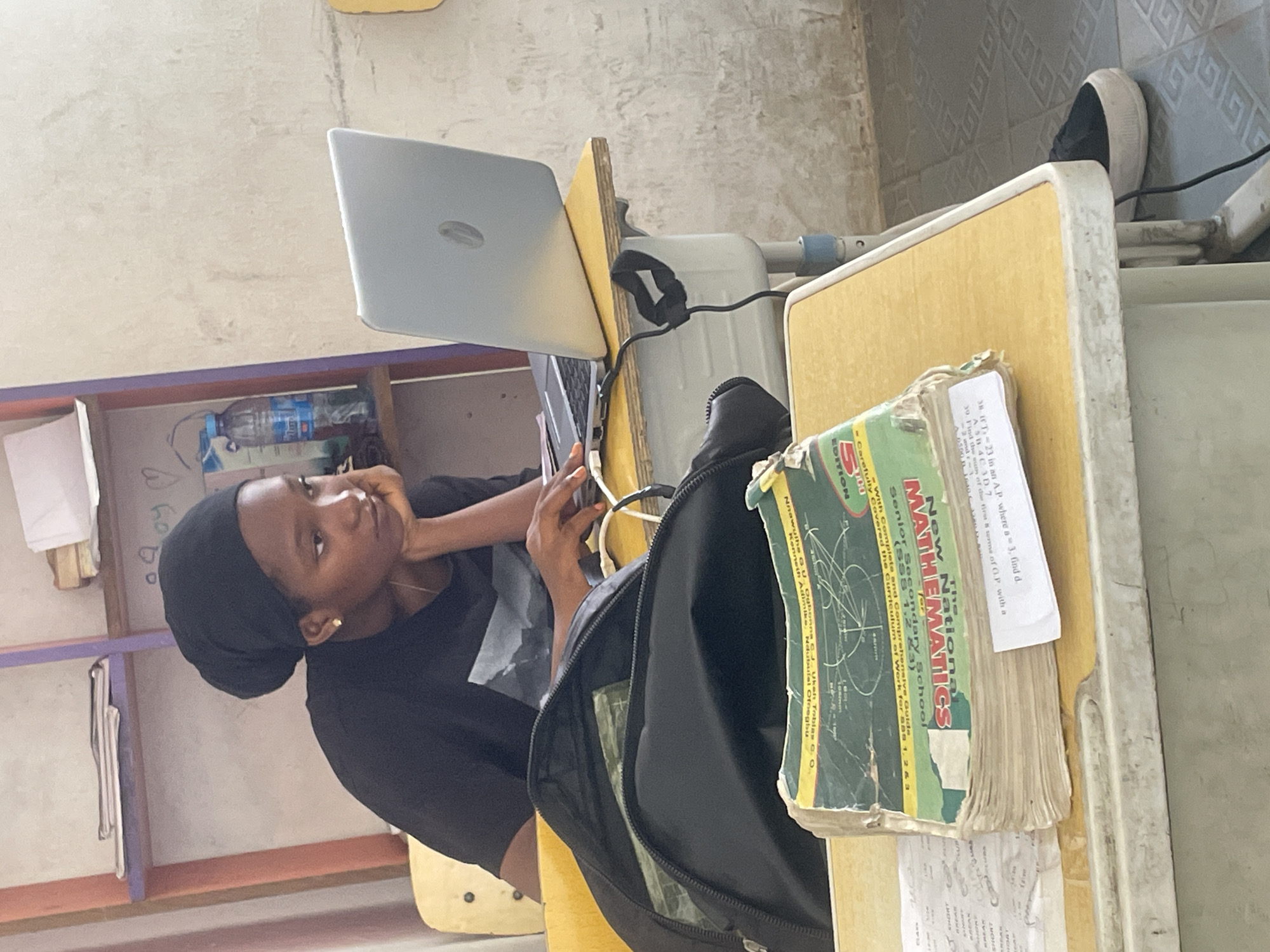 In a classroom in Abuja, Nigeria, a young Black woman wearing a black hijab and black t-shirt is seated at a yellow desk, focused on a silver laptop. Her right elbow rests on the desk, and her chin is propped on her hand as she studies the screen. Beside her on the desk is an open black backpack and a stack of textbooks, the top one titled "New National Senior Secondary MATHEMATICS." A white paper with handwritten text is tucked into the book. A blue plastic water bottle is visible on a shelf in the background, which is decorated with colorful vertical stripes. The walls are a light, textured color, and the floor is tiled with a geometric pattern. The overall scene depicts a student diligently engaged in her studies.