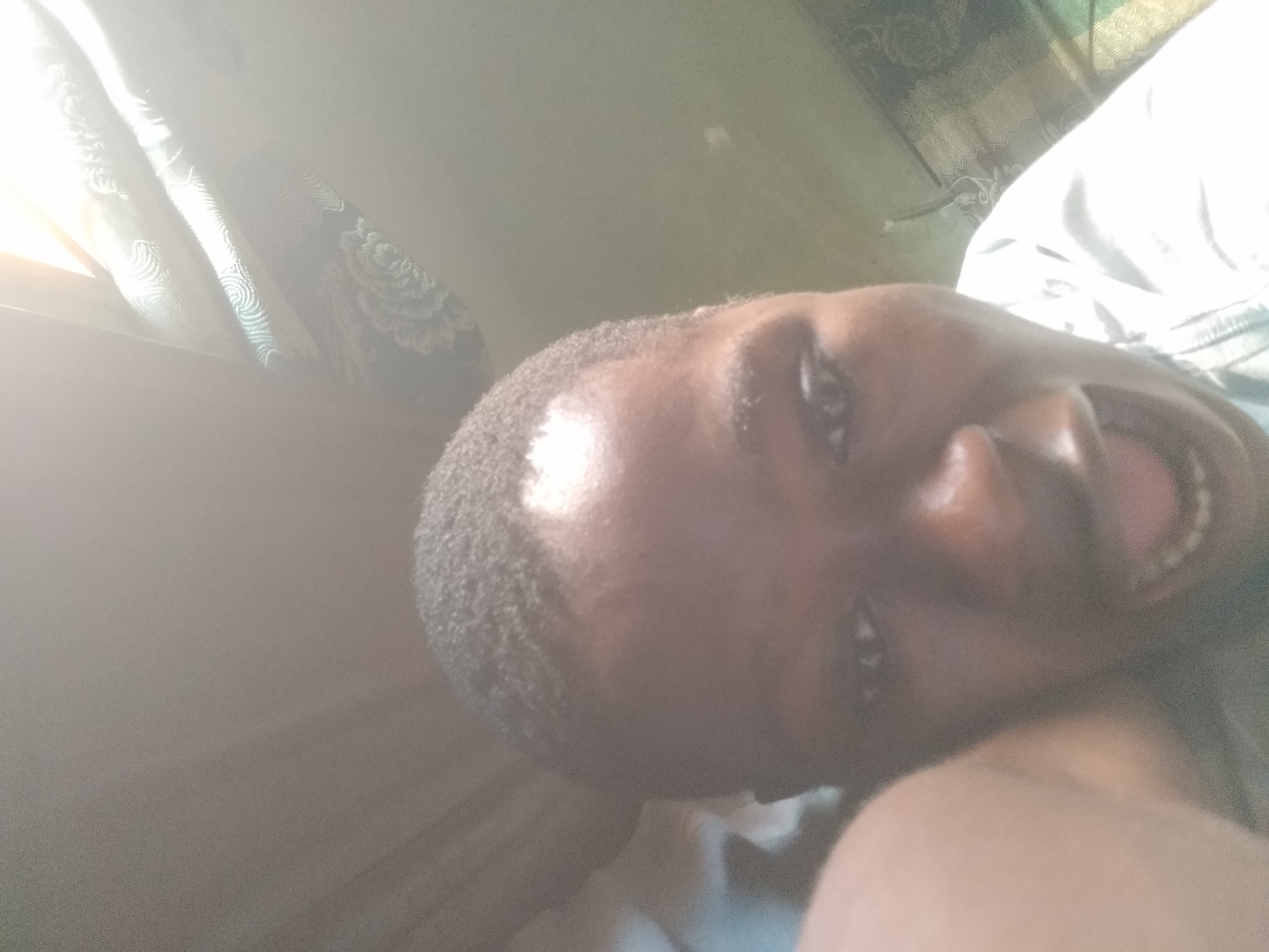 A close-up, tilted selfie of a person with dark skin and short, dark hair. Their mouth is open, revealing teeth and tongue. The person's right shoulder is visible in the lower right corner of the frame. The background shows a window with curtains, partially obscured by bright light. The location and city cannot be determined from the image.