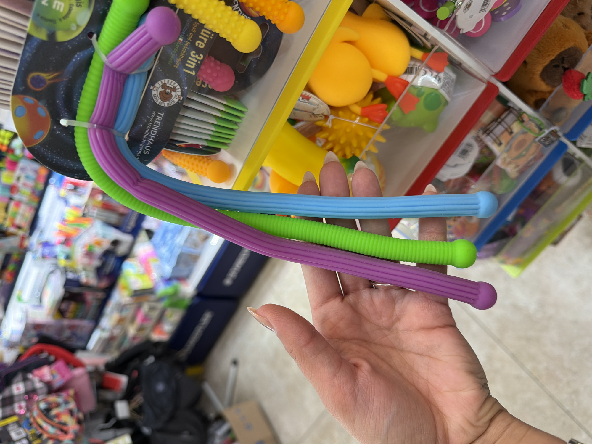 A person's hand holds three colorful, flexible, corrugated tubes (purple, green, and blue) with bulbous ends. The tubes are likely sensory toys. The background features other toys and products displayed in clear plastic containers or shelves. A "TRENDHAUS" package is also visible. Cluttered merchandise including bags are on the floor below the display. The location cannot be determined from the image.
