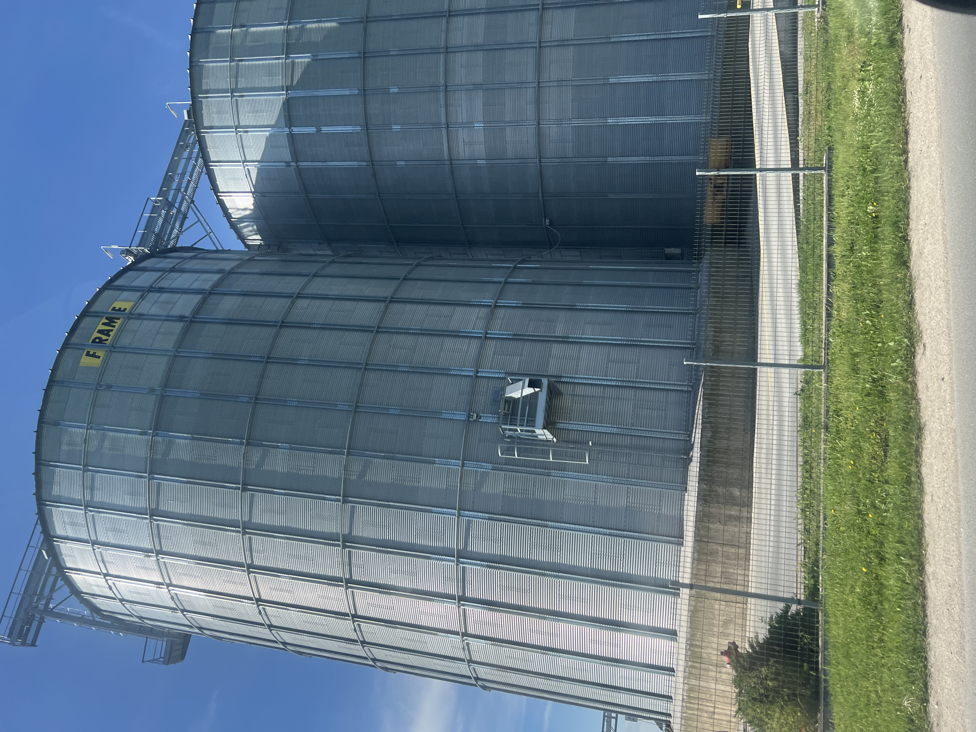 Two large metal grain silos are visible against a blue sky. The silos are cylindrical with corrugated metal siding and a network of metal supports. One silo has the word "FRAME" printed vertically on its side. A metal walkway and ladder system connect the silos. The silos are bordered by a chain-link fence, a concrete or paved area, and a grassy strip with some wildflowers. Beyond the grass is a section of pavement. The scene appears to be in an agricultural or industrial area, potentially in the city of Brezovo, Bulgaria. There are no people visible.