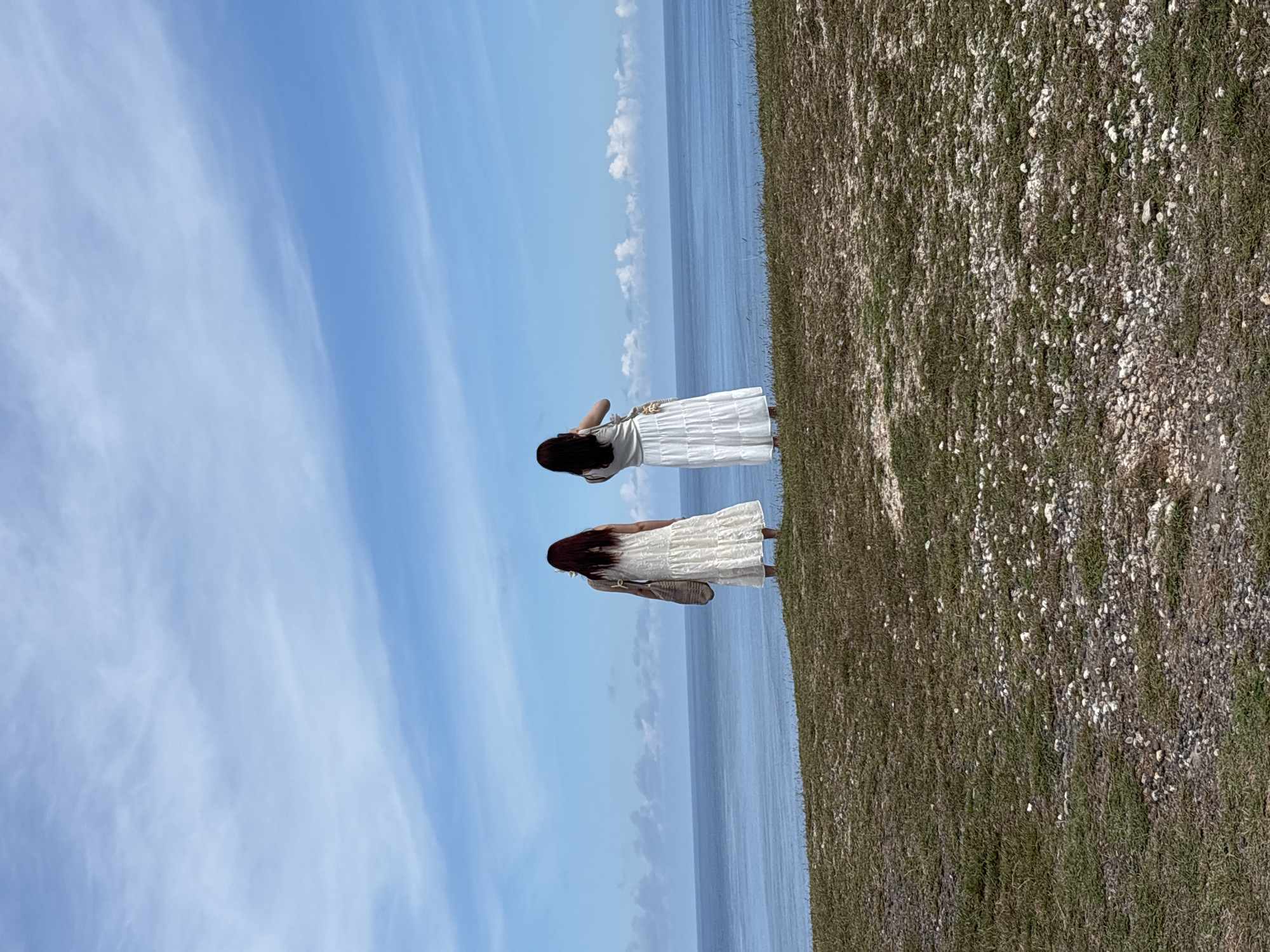 Two women in white dresses stand on a grassy, rocky cliff overlooking the ocean in Kuta Selatan, Indonesia. The sky is blue with wispy clouds. One woman raises her hand to her face, possibly shielding her eyes or adjusting her hair. The other woman carries a woven bag. The scene is peaceful and picturesque.