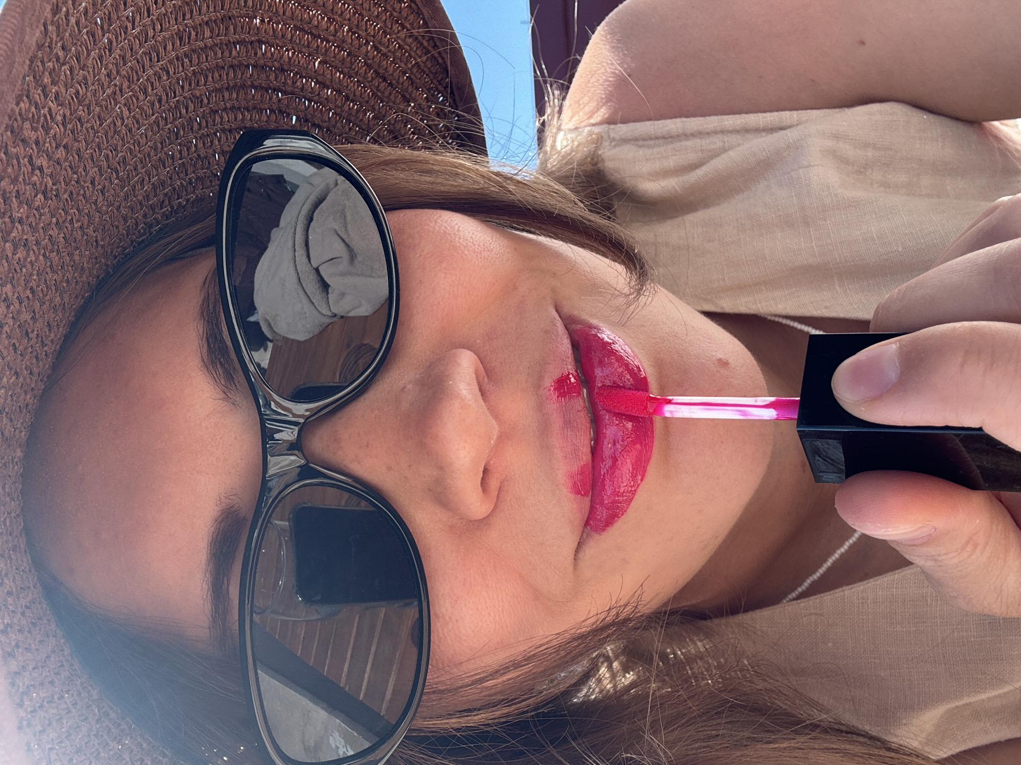 A close-up, angled forensic image shows an individual applying bright fuchsia liquid lipstick in Fethiye, Turkey. The subject wears a wide-brimmed brown straw hat and dark, oval-shaped sunglasses. Reflections in the sunglasses display horizontal wooden slats and a crumpled grey textile. The lipstick is partially applied to the subject's lips and smudged above the upper lip. A pink-tipped applicator, held by the subject's right hand, is positioned at the mouth. The individual wears a light beige sleeveless garment, and a delicate pearl necklace is visible around the neck. A clear blue sky is visible in the background on the right. The scene is brightly lit by natural sunlight.