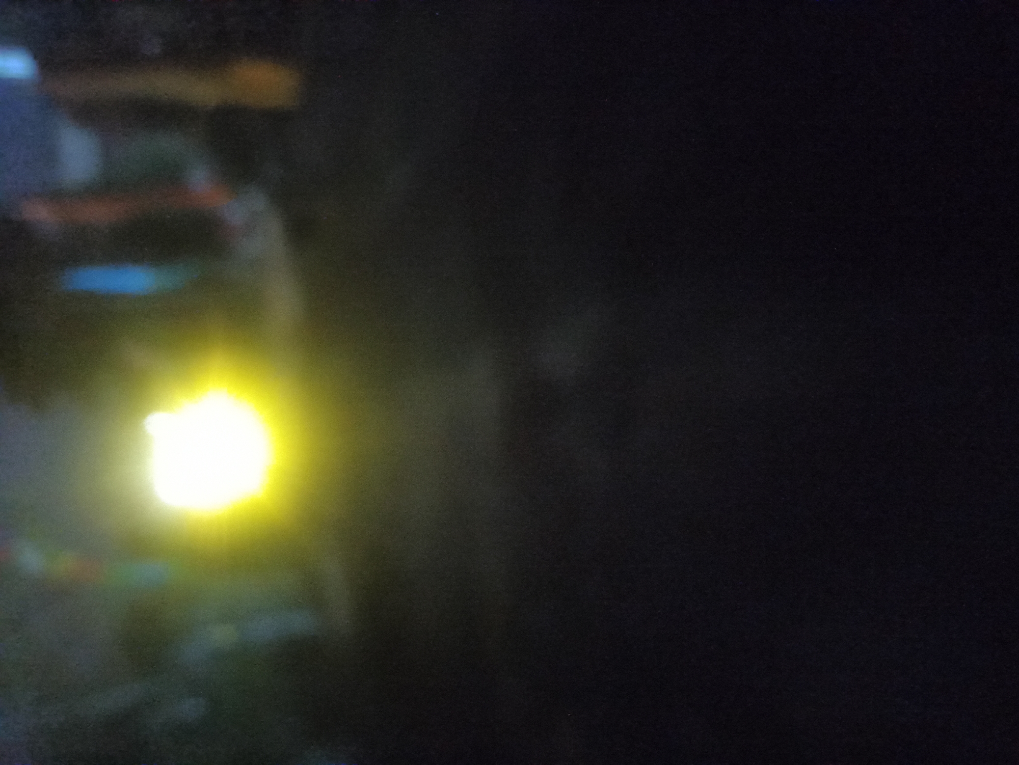 The image is a dimly lit, out-of-focus photograph showing a bright, yellowish light source on the left side. The background is dark and indistinct, with some blurry vertical shapes that appear to be of various colors, including blue and orange, suggesting some objects or people in the distance. The overall impression is one of low light conditions, possibly taken at night or in a dark indoor setting. The image does not provide any specific visual cues or landmarks that would indicate its location in Tuba, Indonesia.