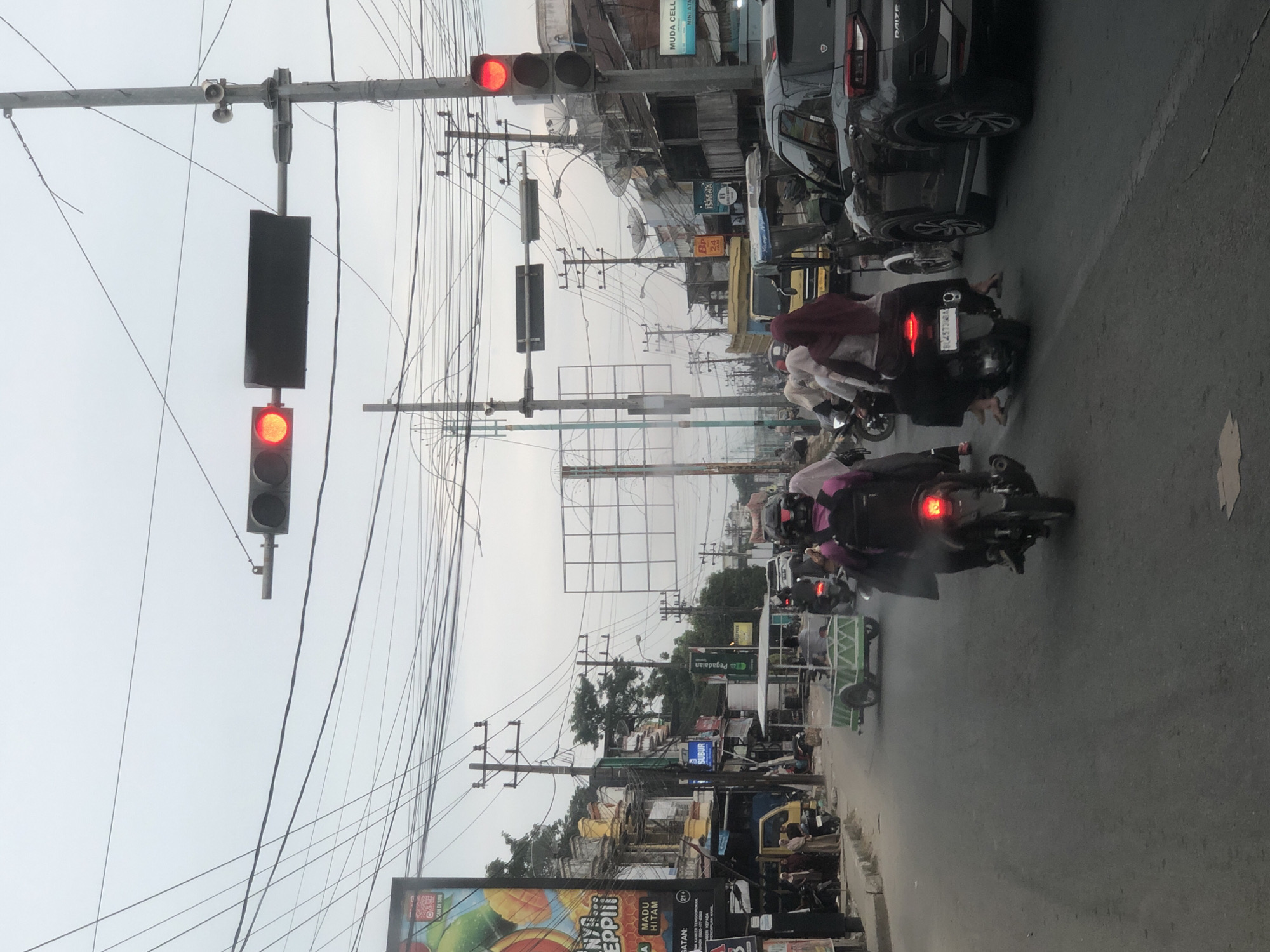The image depicts a street scene in Titibaro, Indonesia, likely during the daytime, with an overcast sky. The foreground shows a paved road with several vehicles, including motorcycles and cars. A person on a motorcycle, wearing a pink jacket and backpack, is prominently featured in the center, with their brake lights illuminated. To their right, another motorcycle carries two individuals, one wearing a maroon hijab. A black SUV is parked to the right of the road.
In the background, a yellow truck is visible. The street is lined with buildings and storefronts, some of which have advertisements. Power lines are strung across the sky, a common sight in urban Indonesian landscapes. Traffic lights are visible, with one red light illuminated, indicating a stop. A small, green trailer is being pulled by a motorcycle. Pedestrians can be seen on the sidewalk on the left side of the road. The overall atmosphere suggests a busy, active street.