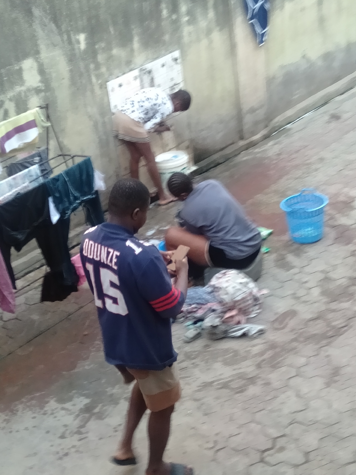 The image depicts an outdoor domestic scene in Shangisha, Nigeria, likely in a backyard or courtyard, characterized by a paved, wet hexagonal tiled ground and a rough, light-colored concrete wall in the background.
Three individuals are present, engaged in laundry-related activities. In the foreground, a person is seen from behind, wearing a blue jersey with the name "ODUNZE" and the number "15" printed in white on the back. They are holding a small, rectangular object, possibly a smartphone, in their hands and are wearing light brown shorts and dark slippers.
In the middle ground, another person is seated, bent over, wearing a grey t-shirt and black shorts. A pile of clothes is visible in front of them on the ground, and a bright blue plastic bucket, possibly containing water or suds, sits nearby. This individual appears to be handwashing clothes. Further in the background, a third person, wearing a white patterned shirt and light brown shorts, is also bent over near a white bucket, indicating their involvement in similar tasks.
To the left of the frame, a metal clothes drying rack is laden with various garments, including a striped towel and dark clothing, suggesting that laundry is an ongoing activity. The ground is noticeably damp and stained in several areas, consistent with water spillage from washing. The lighting is diffused, suggesting daytime under an overcast sky or during the softer light of early morning or late afternoon. The scene portrays routine household chores in a communal outdoor setting.