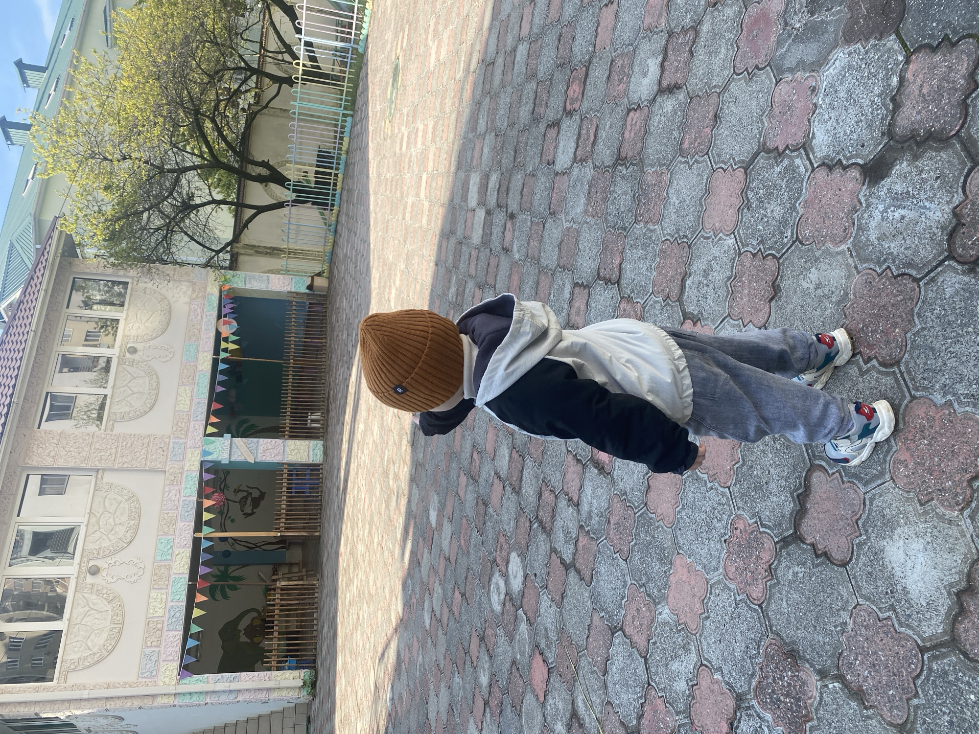 A child wearing a brown knit hat, grey sweatpants, and sneakers is standing on a brick-paved surface outdoors. The child is wearing a jacket with white sleeves and a black body. Behind the child is a building with multiple windows and decorations, including colorful flags. There is a tree to the left of the building. The scene appears to be in Sochi, Russia.