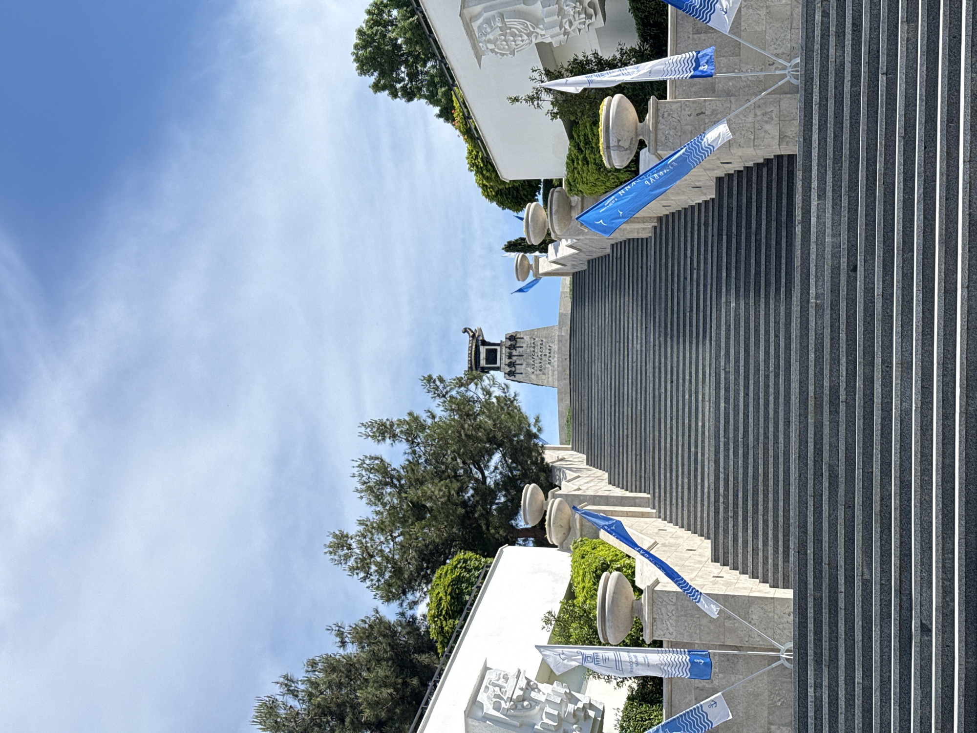 A wide, long staircase made of grey stone ascends towards a monument topped with a structure resembling a traditional East Asian roof. The staircase is flanked by white stone balustrades and urns, with lush green trees and bushes growing on either side and behind the balustrades. Several flags, predominantly white with blue wave patterns and anchors, and solid blue flags, are displayed on poles along the sides of the staircase. The sky above is a bright blue with sparse white clouds. The scene appears to be a monument or memorial site in an urban environment.