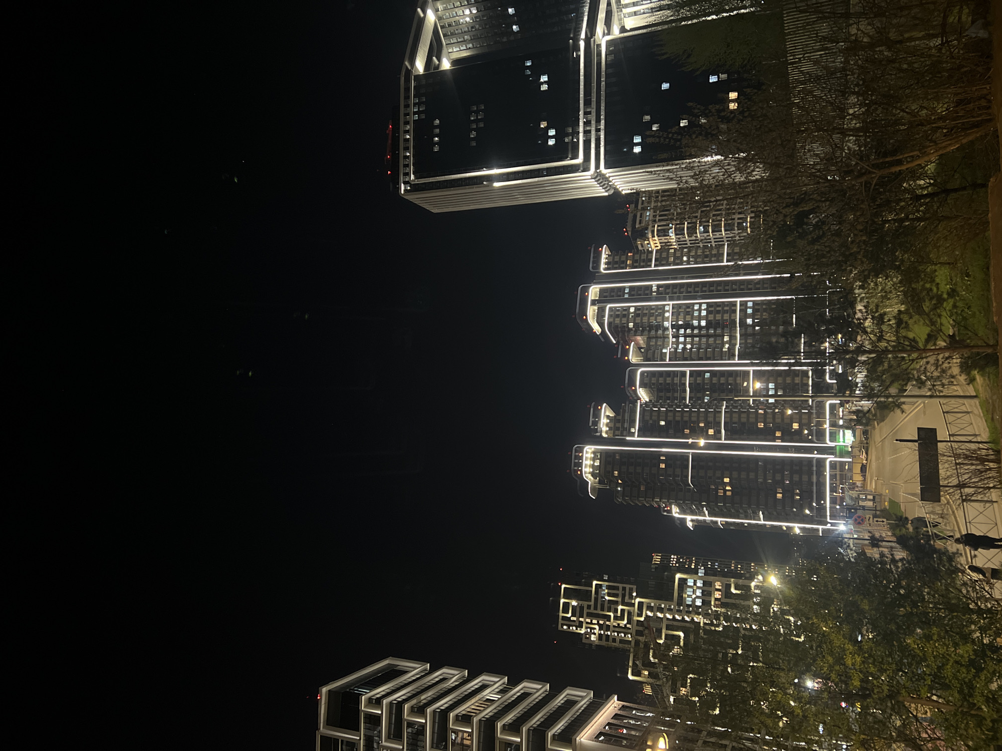 The image shows a nighttime view of illuminated buildings in Moscow, Russia. The buildings are modern high-rises with distinct architectural designs, featuring linear lighting accents. Trees are visible in the foreground, slightly obscuring the view of some buildings. A street with a traffic sign is partially visible on the right side of the image. A silhouette of a person is partially visible along the street. The sky is dark and clear.