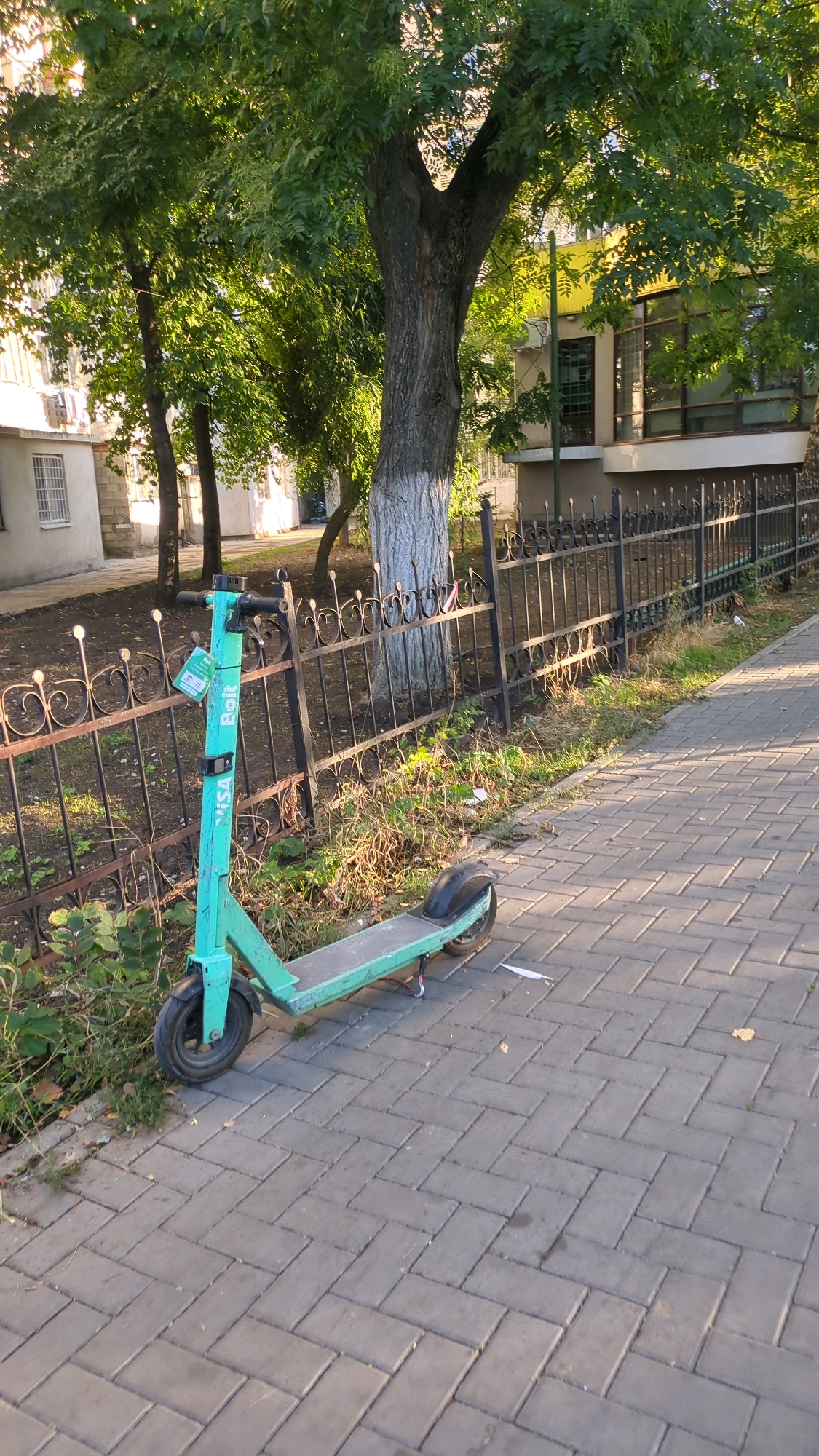 An electric scooter, identified as a Bolt scooter by its branding, is parked on a paved sidewalk next to a black metal fence. The scooter is teal in color and appears to be in a slightly weathered condition. Behind the fence, there is an area with dry grass and some debris. Further back, several trees with green foliage are visible, casting shadows on the scene. A building with windows and a yellow facade is partially visible behind the trees. The overall setting suggests an urban environment, likely in Chișinău, Moldova, given the typical availability of shared electric scooter services in such cities. The lighting indicates it is daytime, possibly late afternoon or early morning due to the direction of the shadows. There are no people or animals present in the frame.